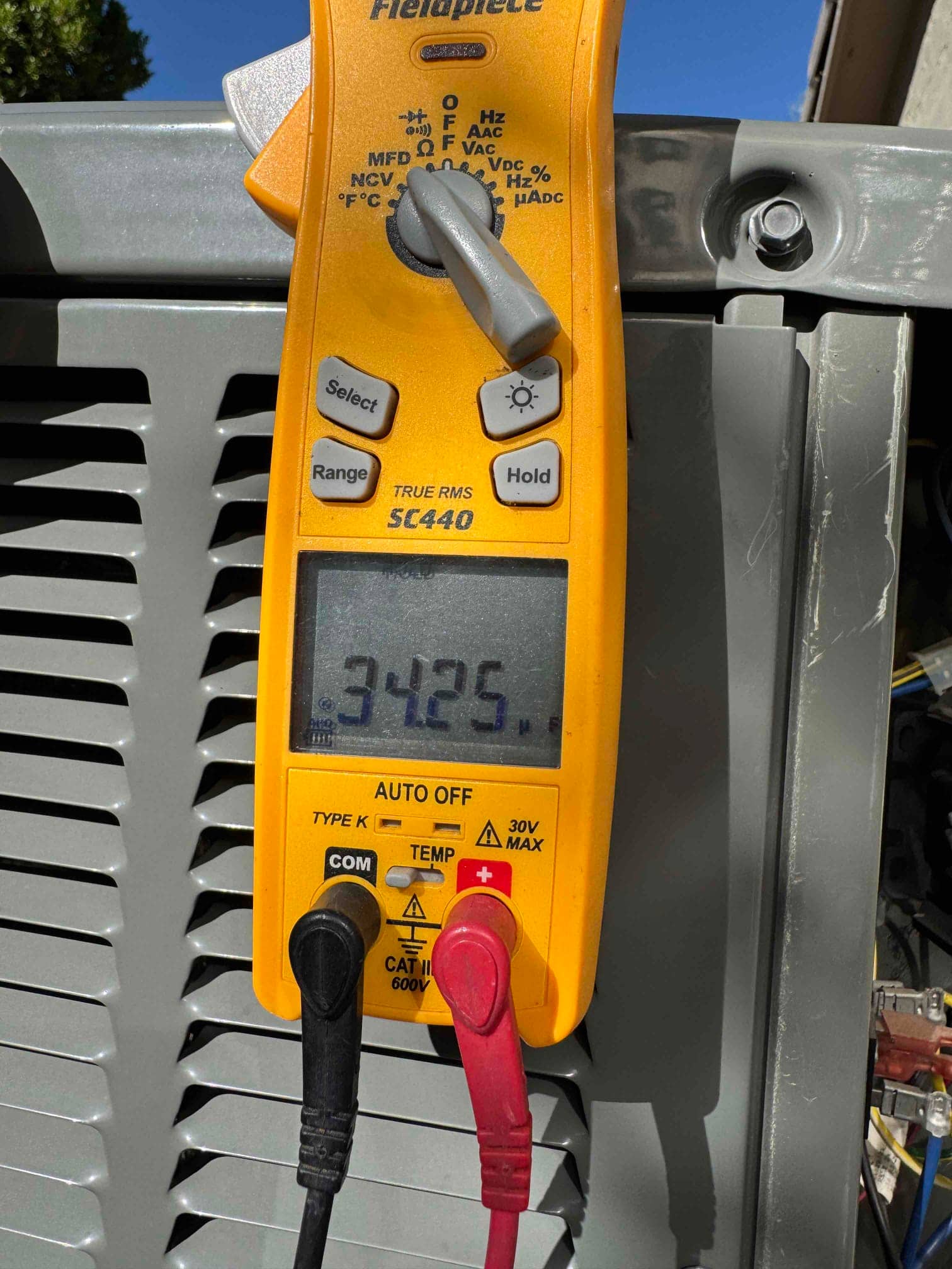 Digital multimeter measures 34.25 μF using red and black leads while testing a capacitor on the outdoor unit.