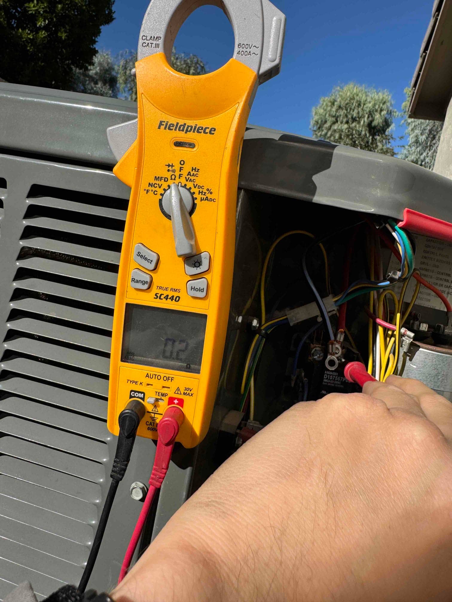 Digital multimeter probes the contactor wiring inside the condenser, displaying 0.2 V on the screen during testing.
