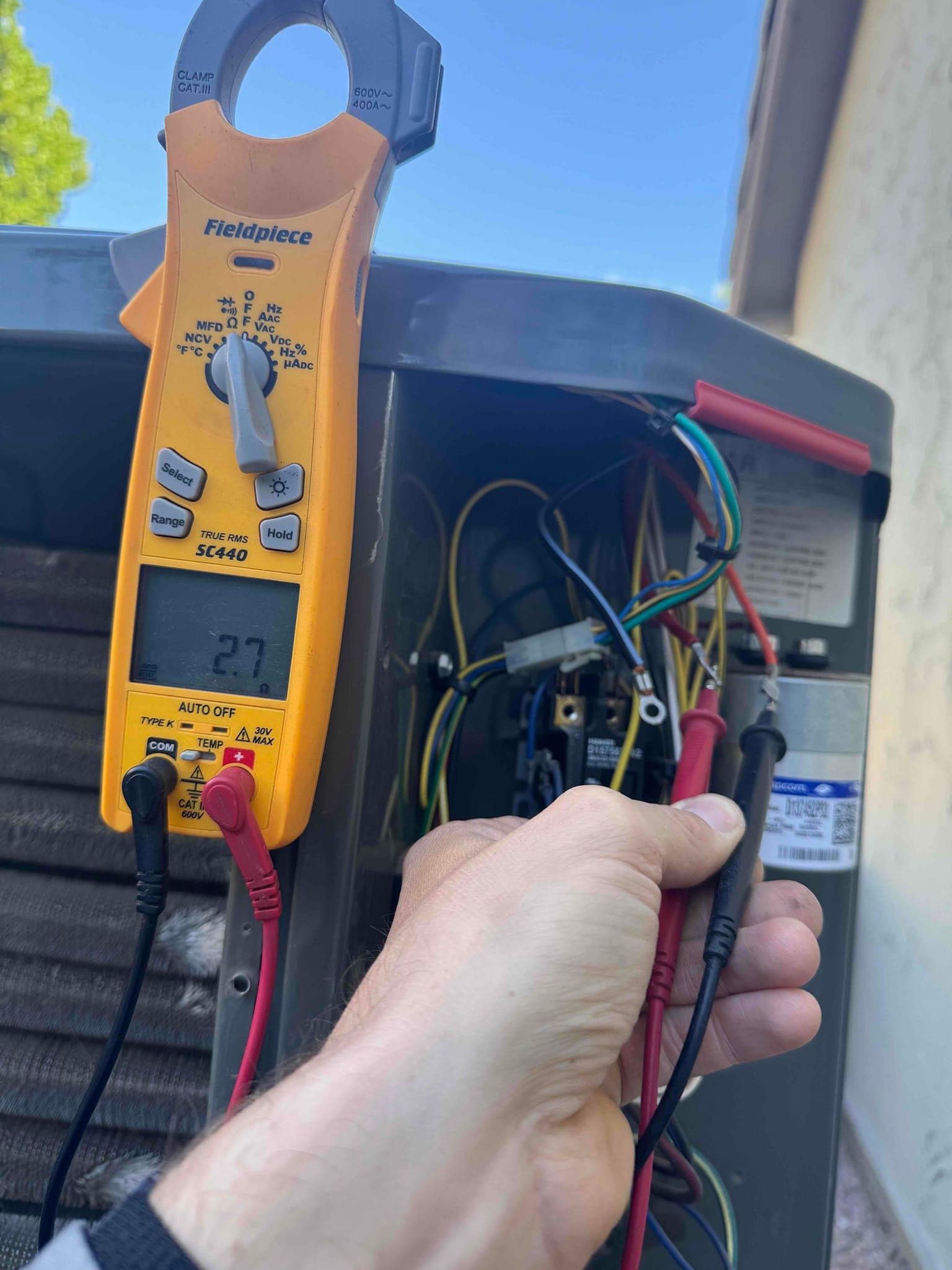 Digital multimeter shows 27.7 V measured across the low-voltage control terminals inside the outdoor condenser unit.