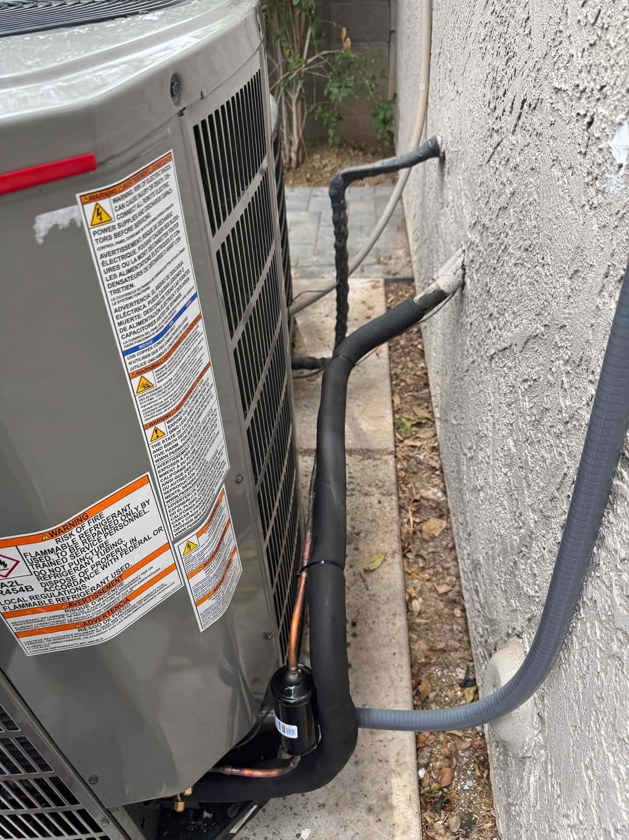 New outdoor condenser set on pad with insulated refrigerant lines, suction accumulator, and electrical whip entering the wall penetration.
