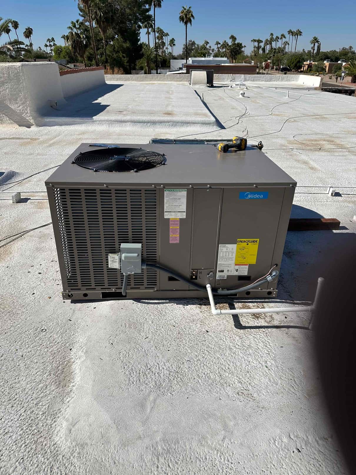 New package HVAC unit set on a flat roof with connected electrical whip, disconnect, and condensate drain piping visible.