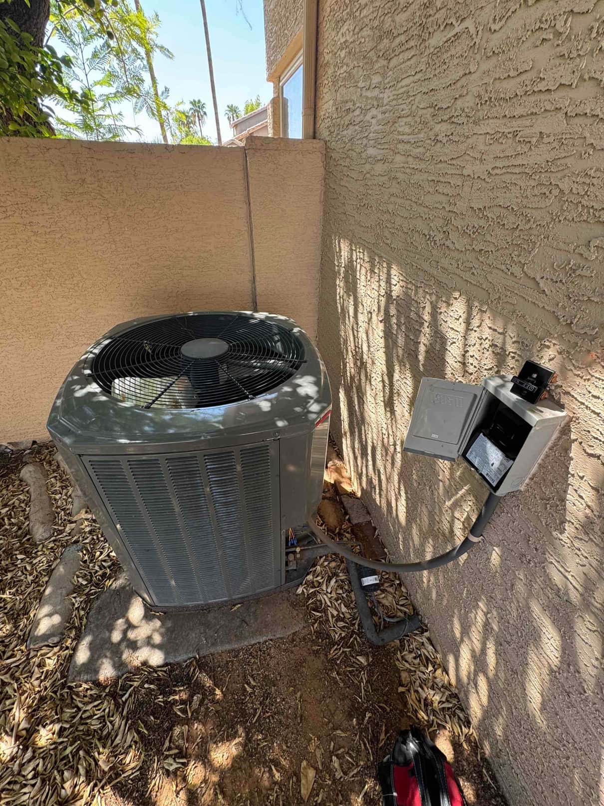 Outdoor air conditioning condenser sits beside a stucco wall with disconnect box and whip conduit installed.