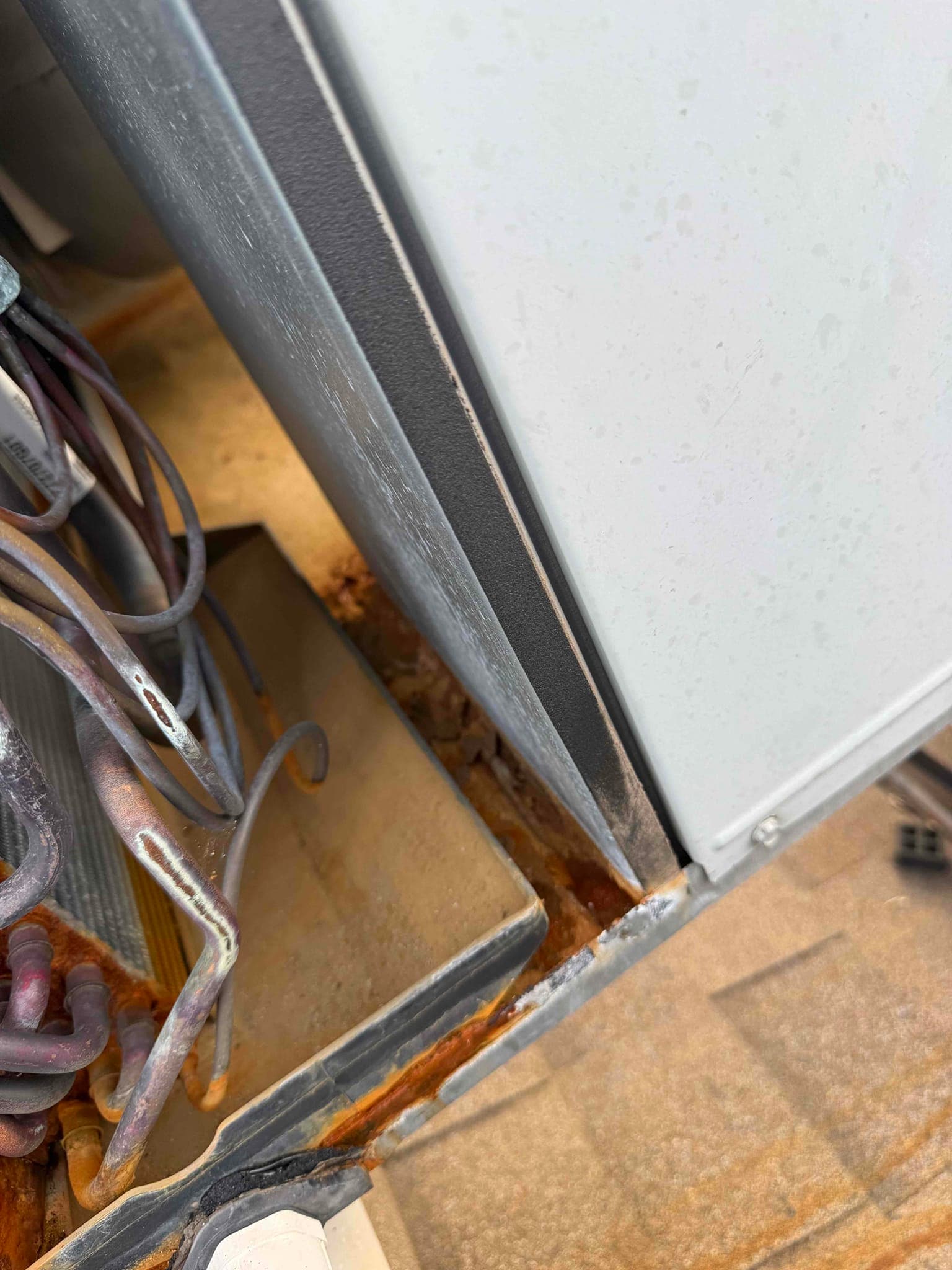 Outdoor condenser cabinet partially open, exposing corroded base pan, rusted edge, and wiring near the coil section.