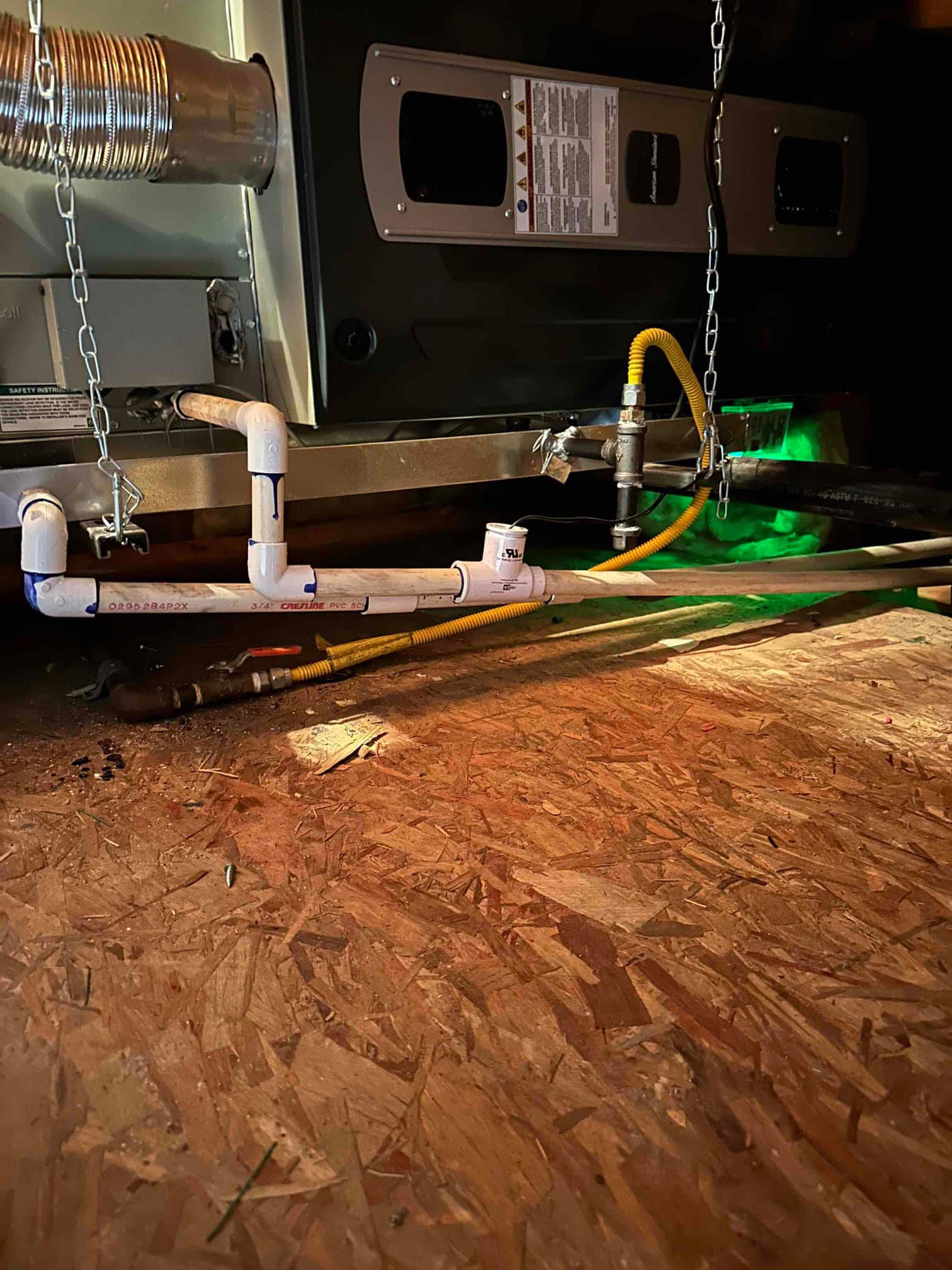 Suspended air handler installation with PVC condensate drain, gas flex connector, and vent piping secured over a plywood crawlspace floor.