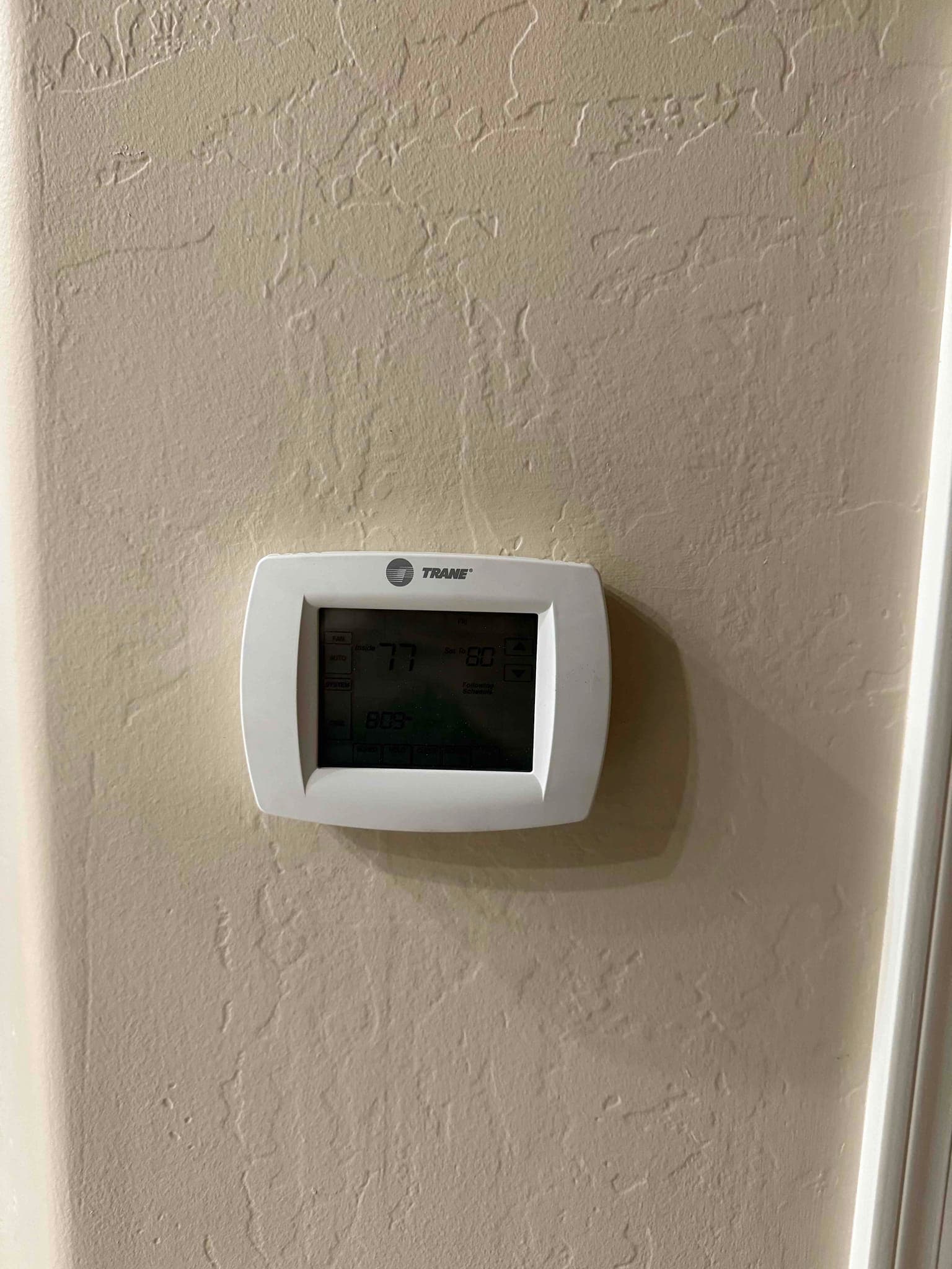 Wall-mounted digital thermostat displays indoor temperature and operating mode on a lighted screen during an air conditioning service visit.