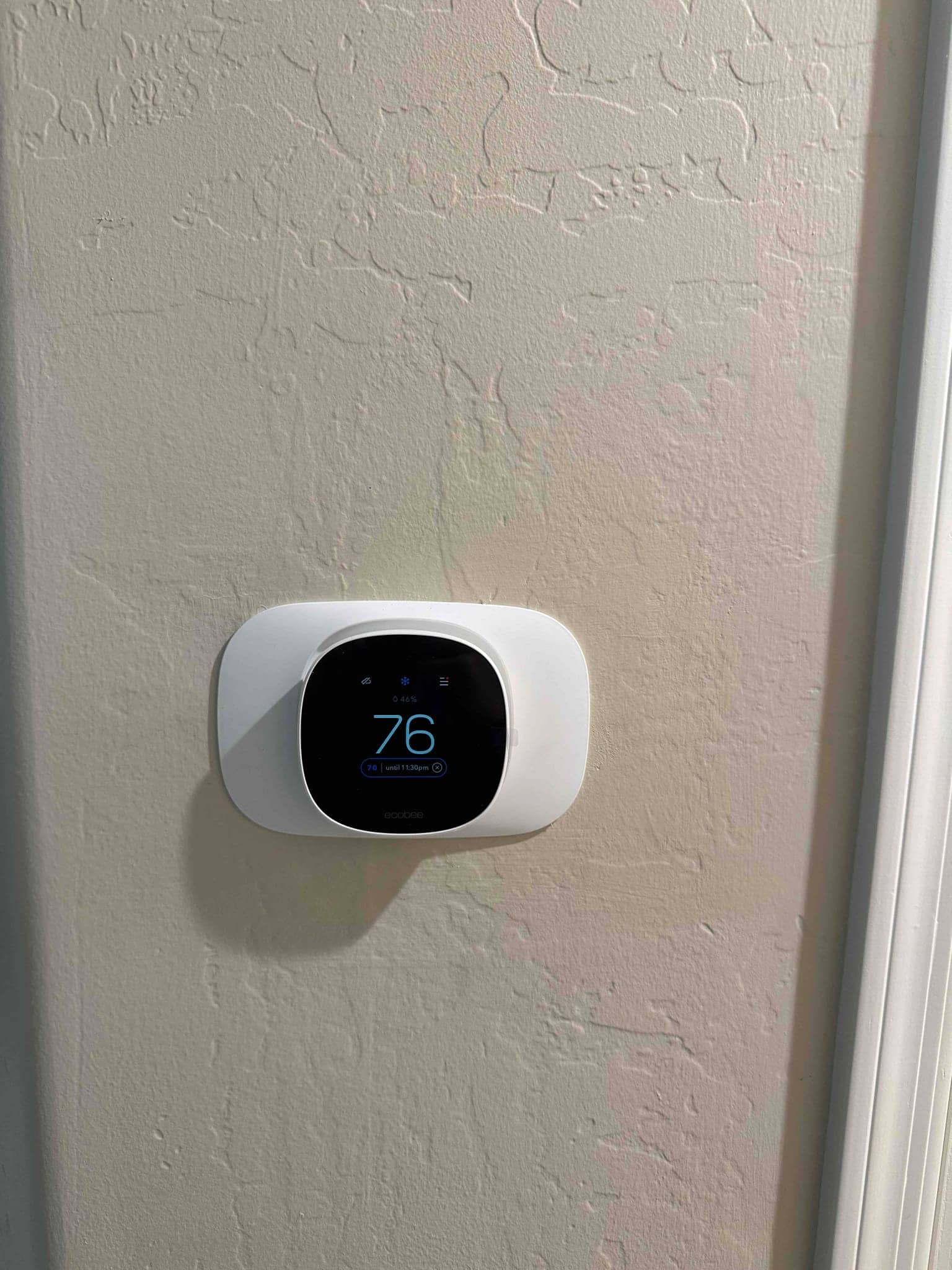 Wall-mounted smart thermostat displays 76 degrees on its screen after installation, with clean trim plate against textured wall.