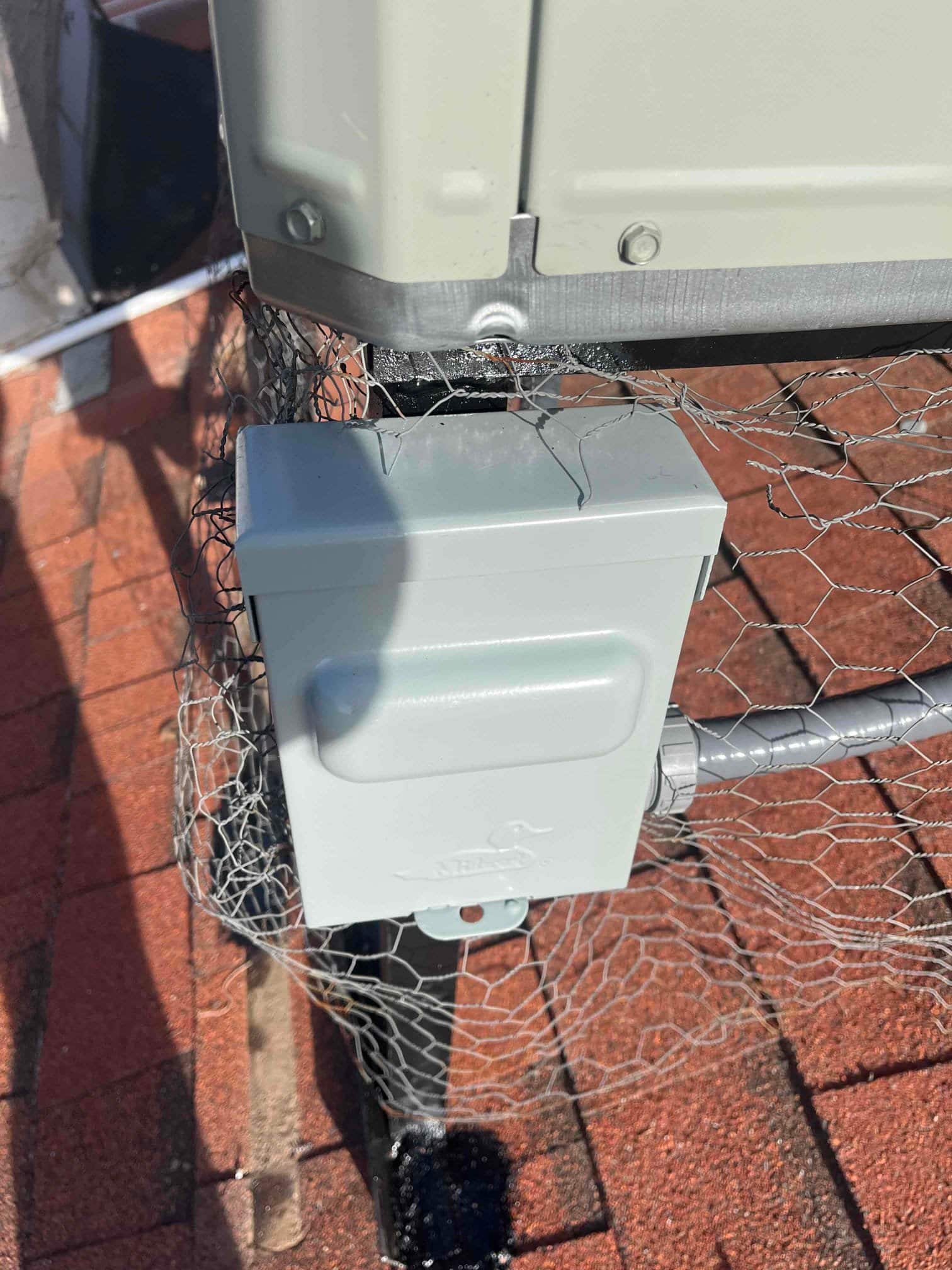 Weatherproof disconnect box mounted beside the outdoor unit with flexible conduit connection and protective wire mesh around the stand.