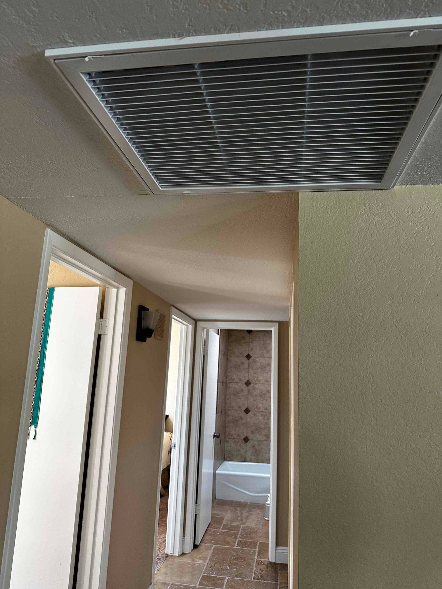 Ceiling-mounted return air grille installed in a hallway near bedroom and bathroom doors with clean, aligned louvers.