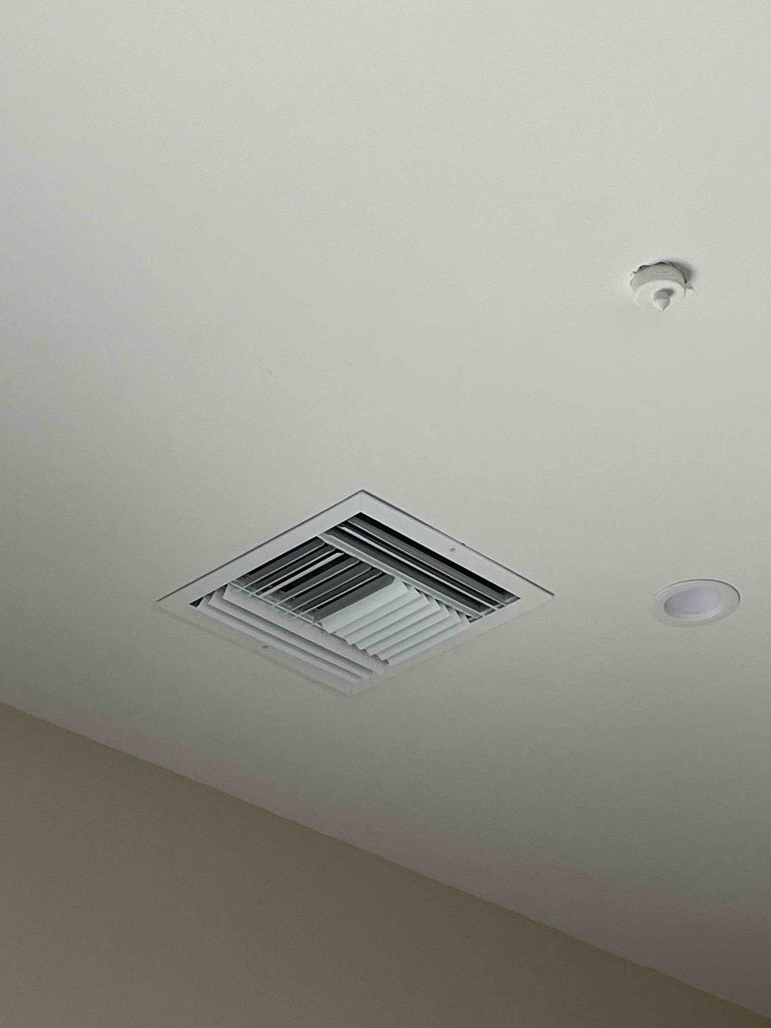Ceiling supply register installed flush with the drywall near a recessed light and sensor on a finished ceiling.