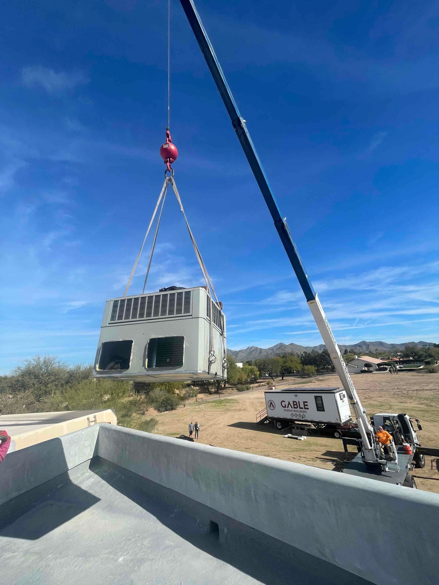 Crane lifts a large rooftop HVAC package unit into position during installation, viewed from the roof near the curb opening.
