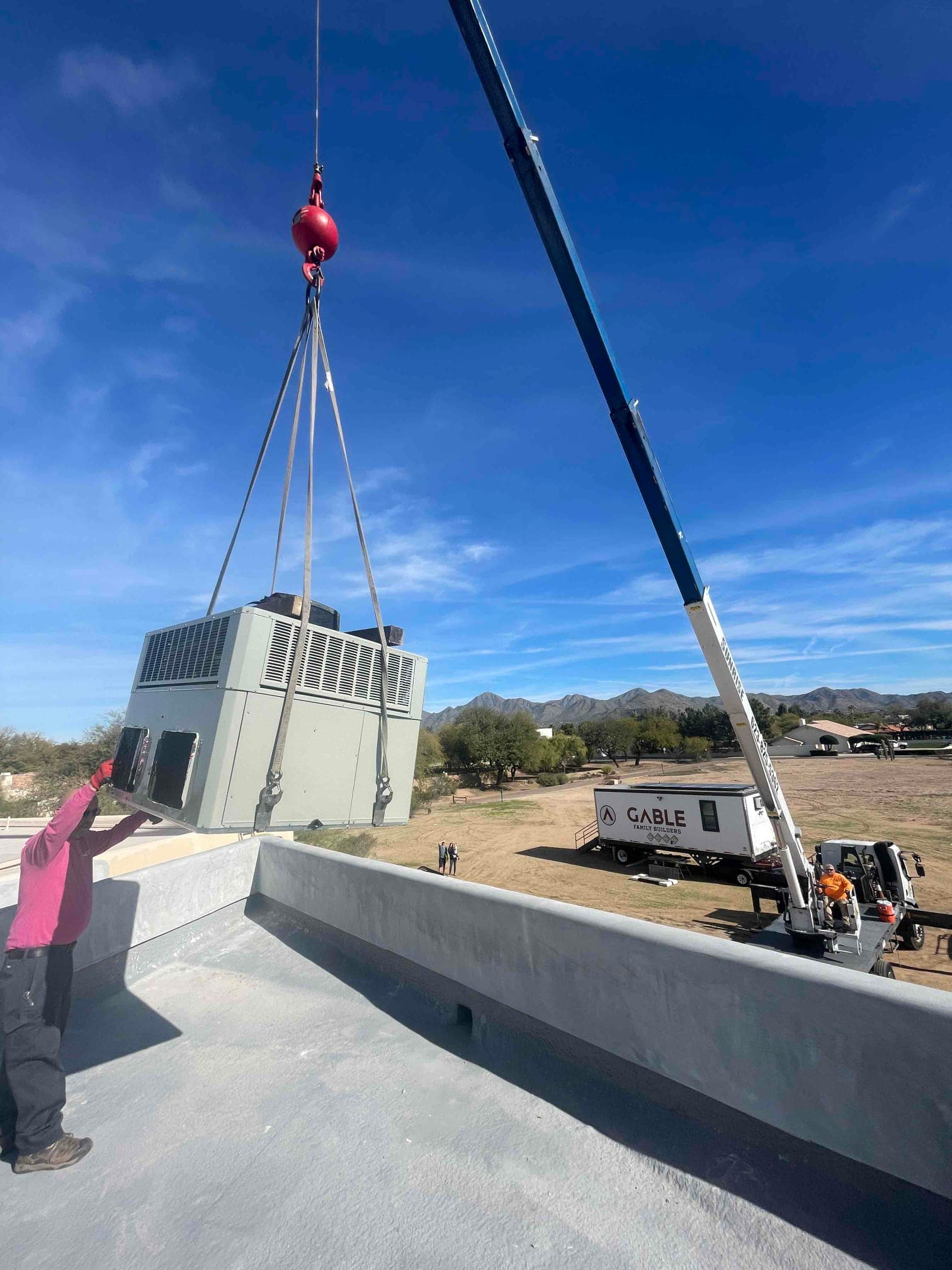 Crane lifts a large rooftop HVAC package unit into position while a technician guides placement on the roof curb.