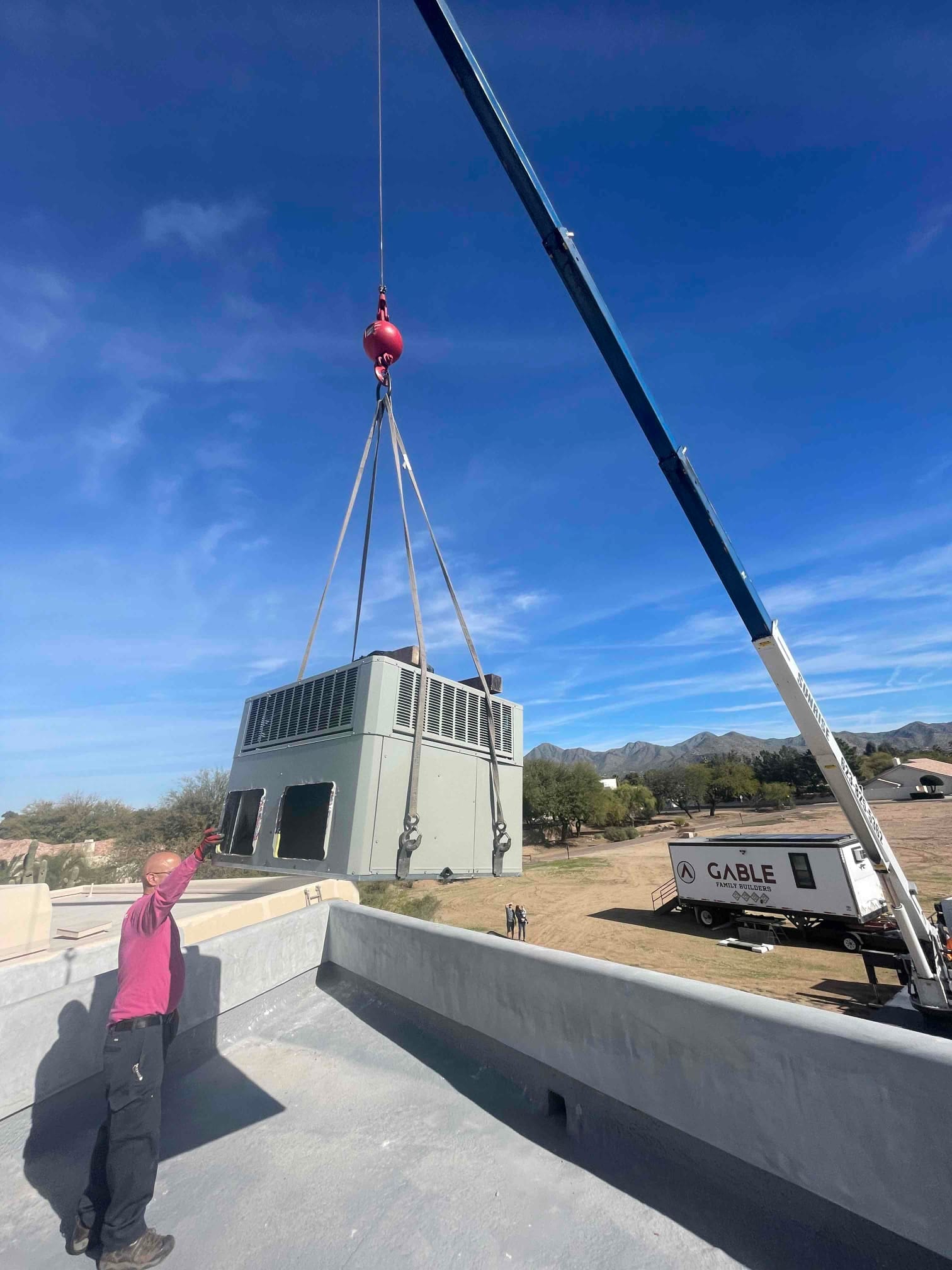 Crane lifts a large rooftop HVAC unit into position while a technician guides the placement from the roof edge.