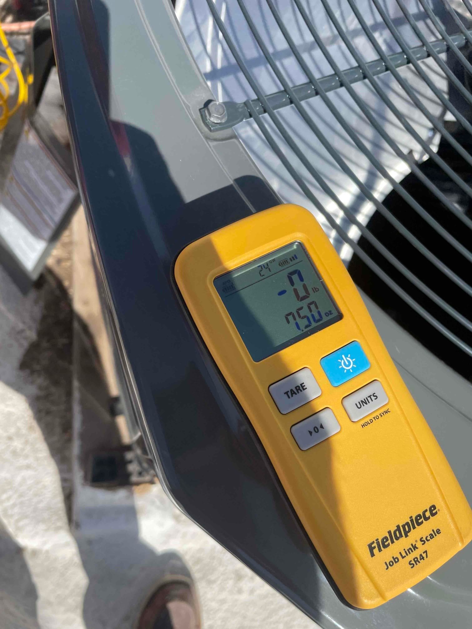 Digital refrigerant scale rests on top of the outdoor condenser during charging setup, showing a zeroed reading and battery status.