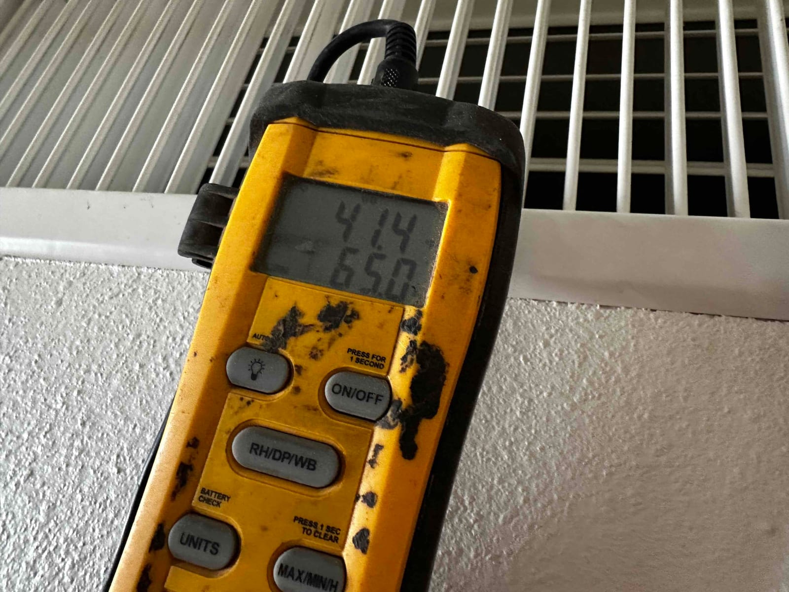 Digital thermometer held at a ceiling grille displays 65.0 degrees Fahrenheit during air temperature check.