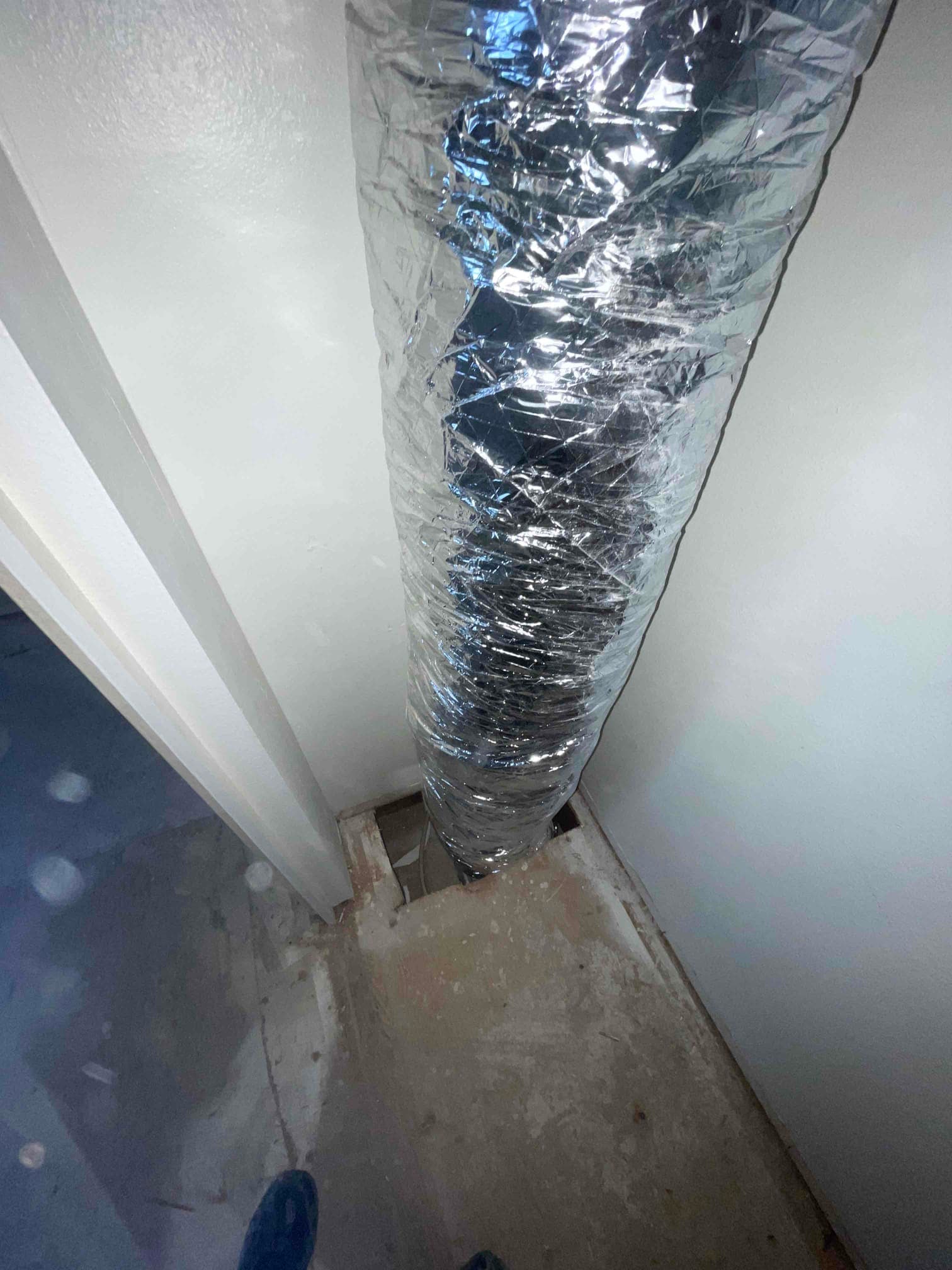 Flexible insulated duct routed through a floor opening during HVAC installation, with surrounding framing and unfinished subfloor visible.