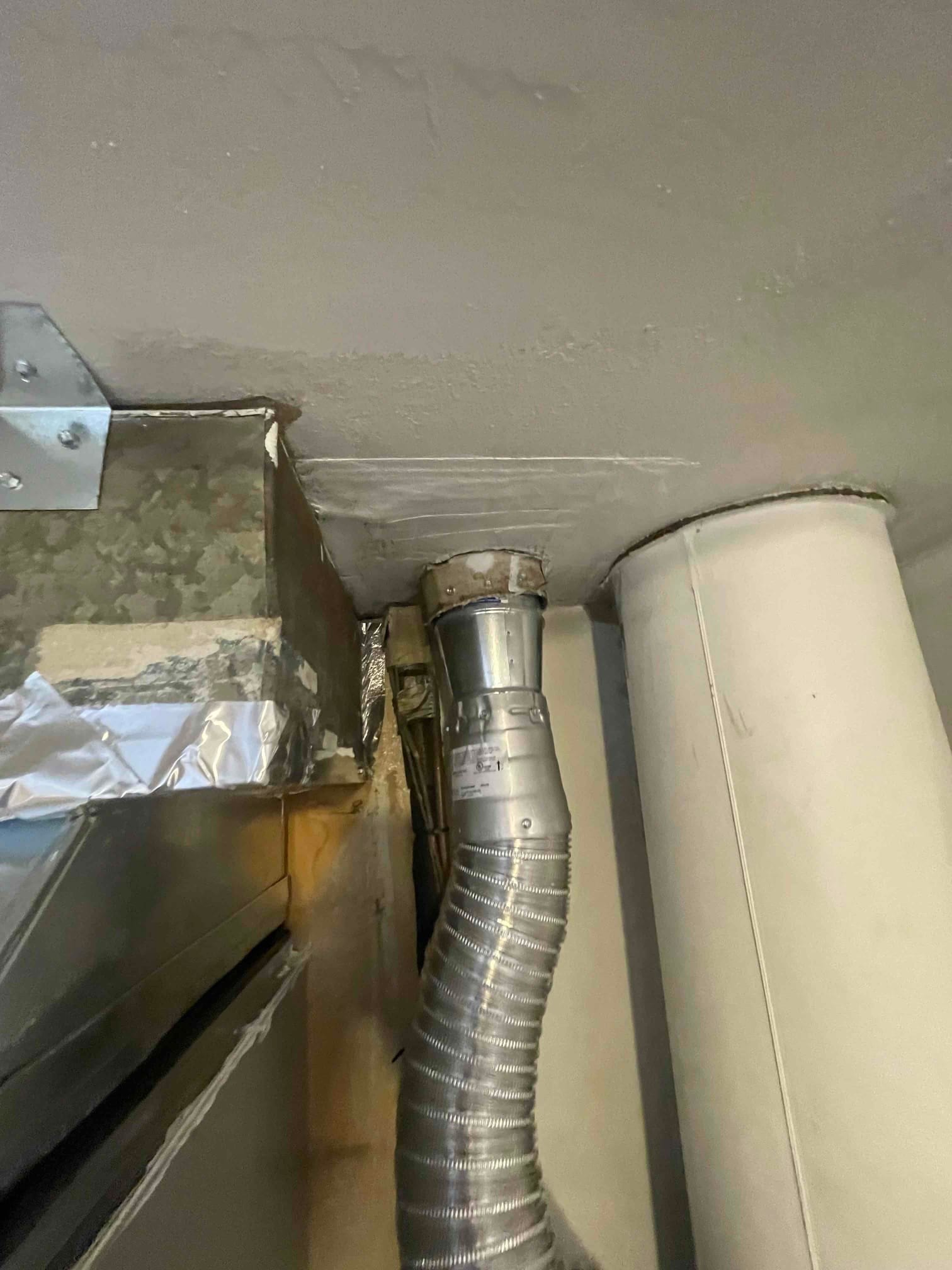 Flexible metal vent connector tied into a vertical flue beside sealed ductwork, showing completed venting near the furnace.