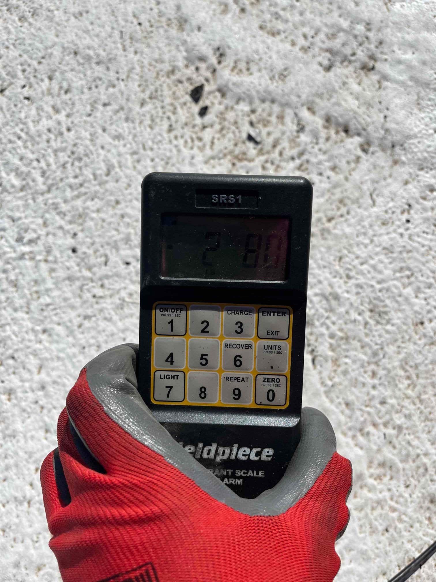 Handheld refrigerant scale controller with digital display showing a partial reading, used during charging of a new system.
