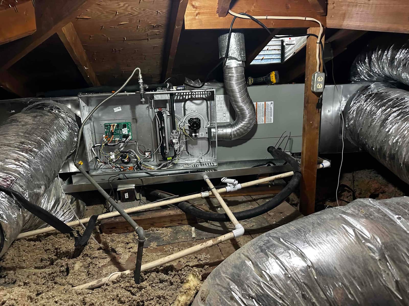 Horizontal attic air handler installed with insulated flex ducts, condensate drains, and access panels open showing internal controls and wiring.