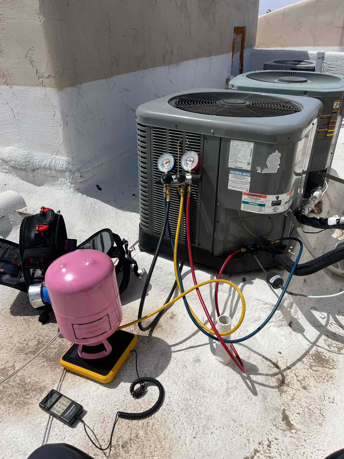 Manifold gauges connected to an outdoor condenser during R-410A charging, with a pink refrigerant cylinder on a digital scale.