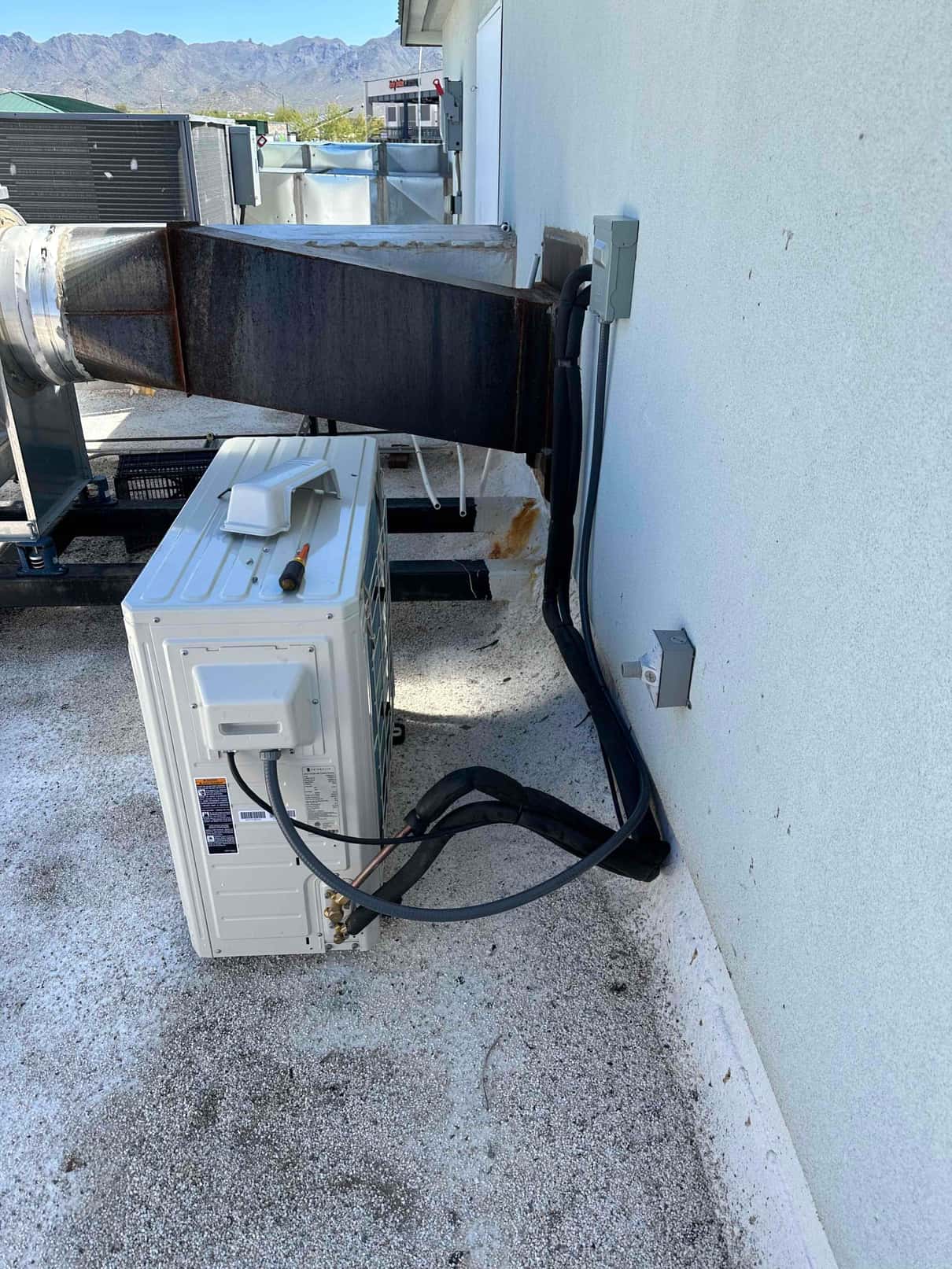 Mini-split condenser set on rooftop with insulated refrigerant lines, electrical disconnects, and wall penetration sealed at the exterior.