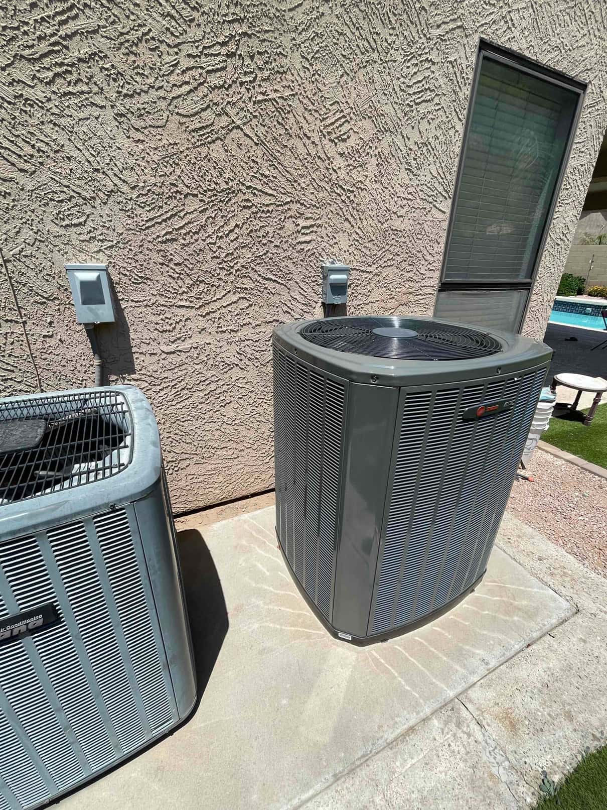 New outdoor heat pump condenser set on a level concrete pad beside an older unit, with weatherproof disconnects mounted on the wall.
