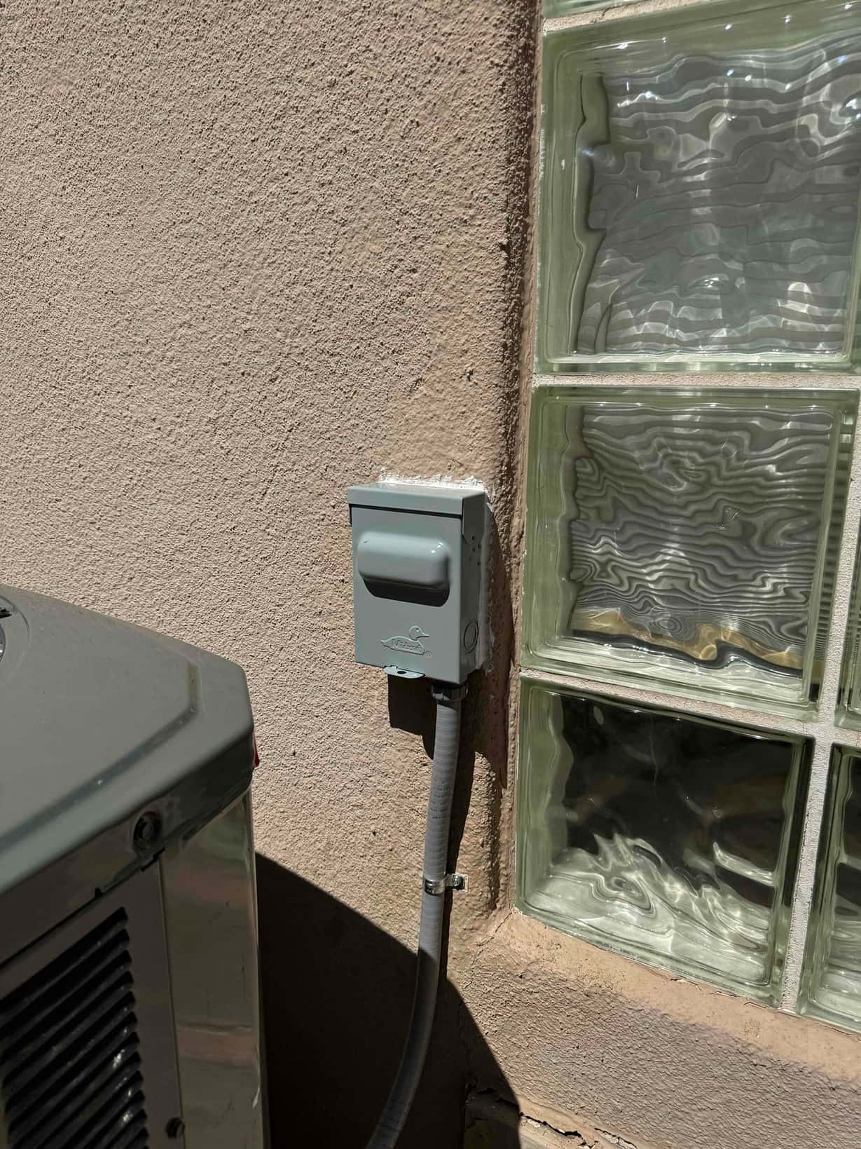 Outdoor AC disconnect box mounted beside the condenser with flexible conduit secured to the stucco wall.