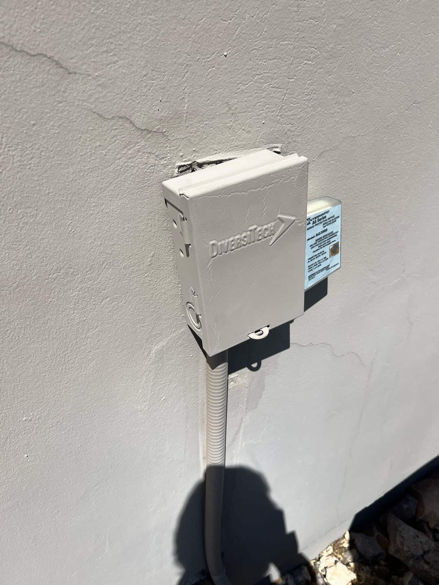 Outdoor AC disconnect box mounted on exterior wall with flexible conduit entering from below and cover closed.