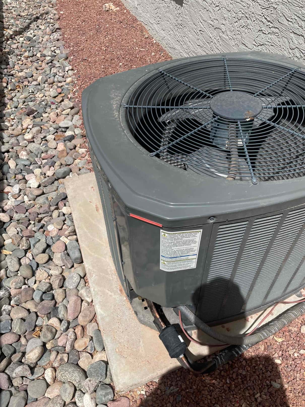 Outdoor air conditioner condenser on a concrete pad with fan grille, coil panels, and insulated refrigerant lines entering the cabinet.