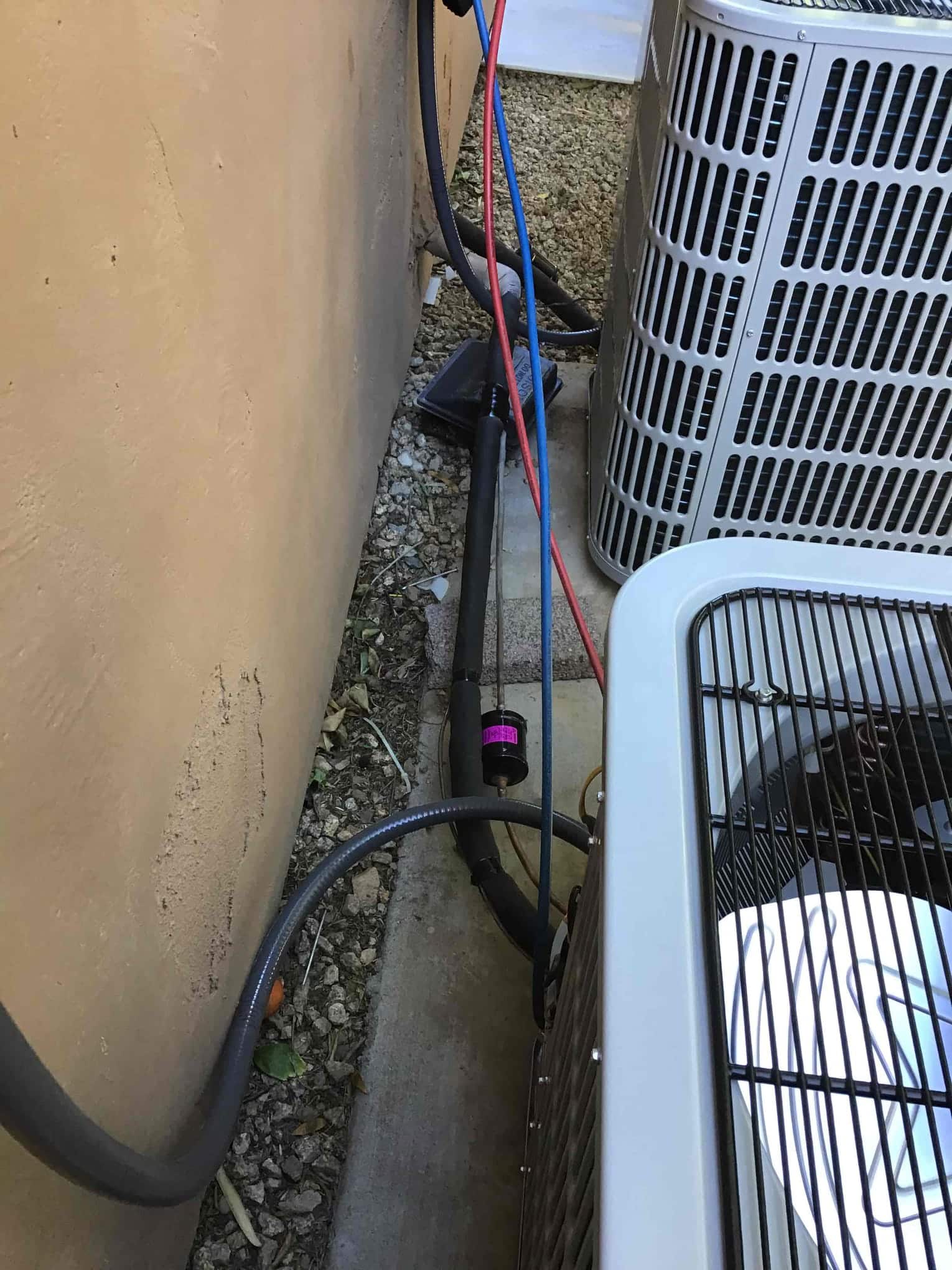 Two outdoor condenser units set on pads with insulated refrigerant lines, electrical whip, and hoses routed along the wall.