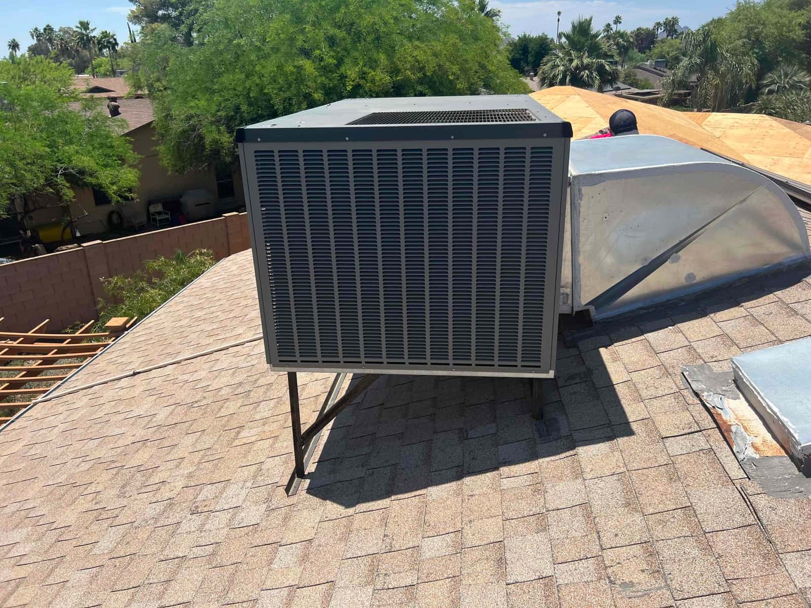 Outdoor condensing unit on a roof stand, next to sheet-metal ductwork and roof curb, under direct sunlight.