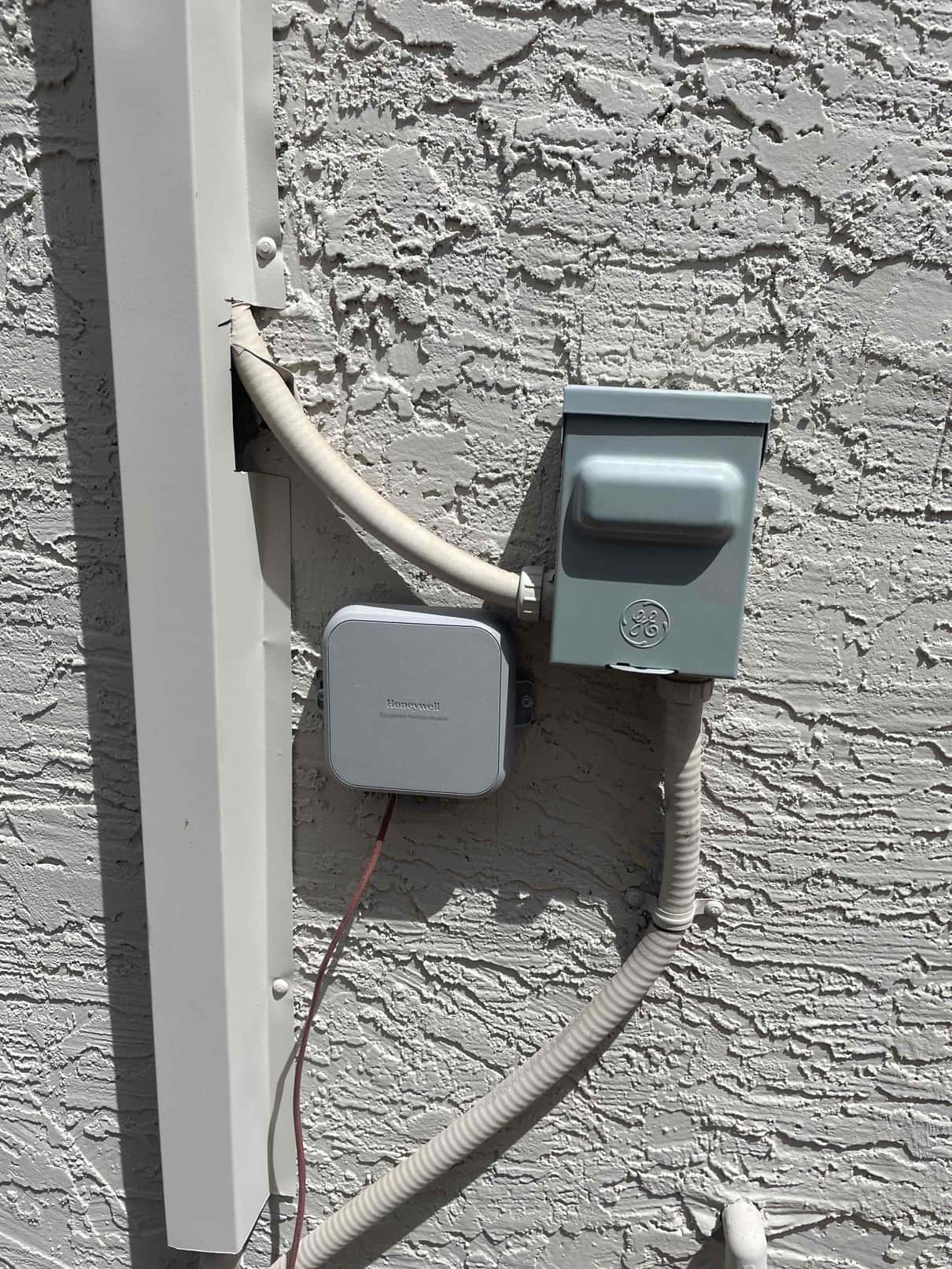 Outdoor electrical disconnect and flexible conduit next to gray control box, with insulated line set exiting wall and a red low-voltage wire visible.
