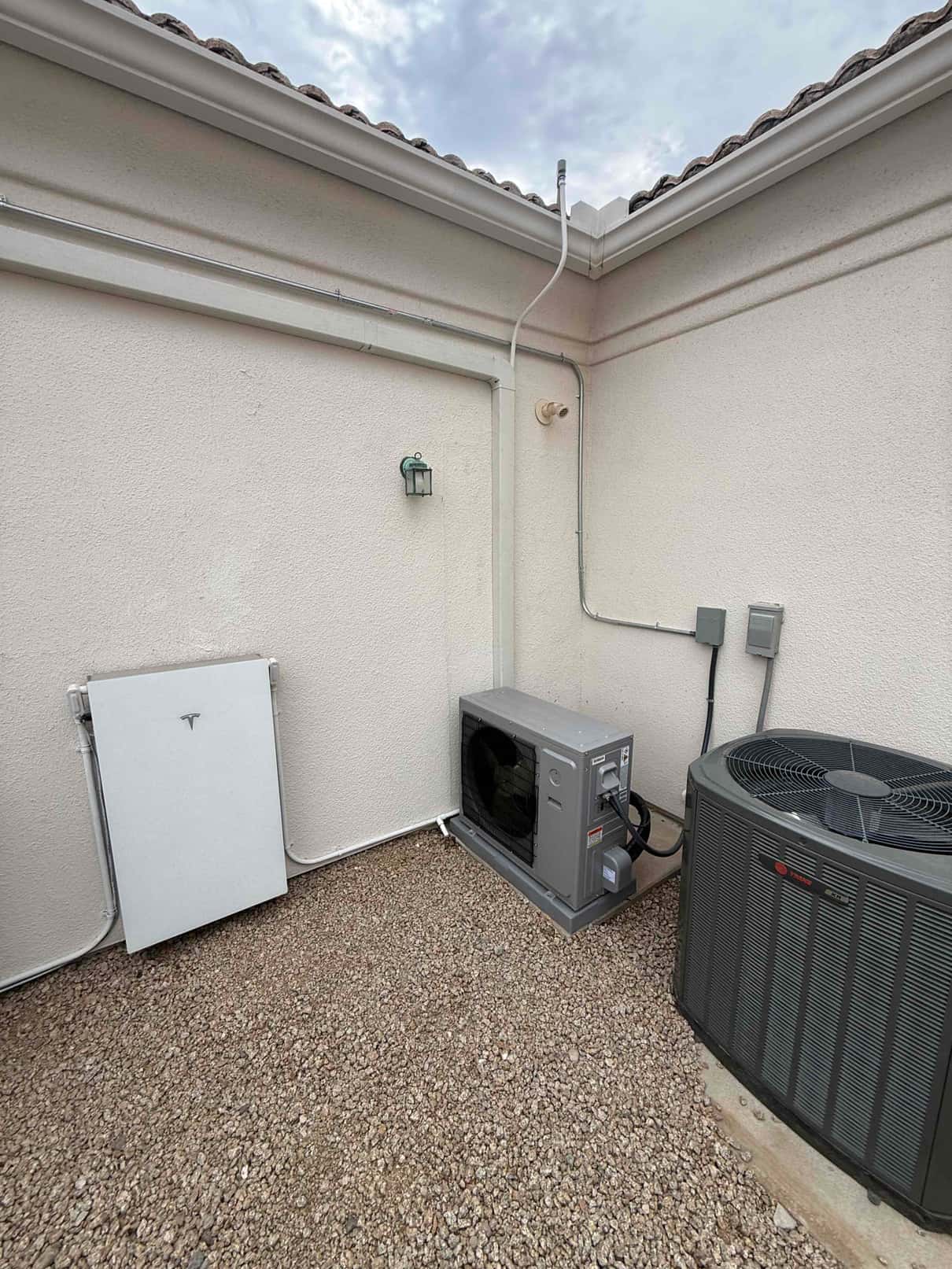 Outdoor installation area showing new mini-split condenser on a pad, adjacent central condenser, electrical disconnects, and properly routed line set conduit.