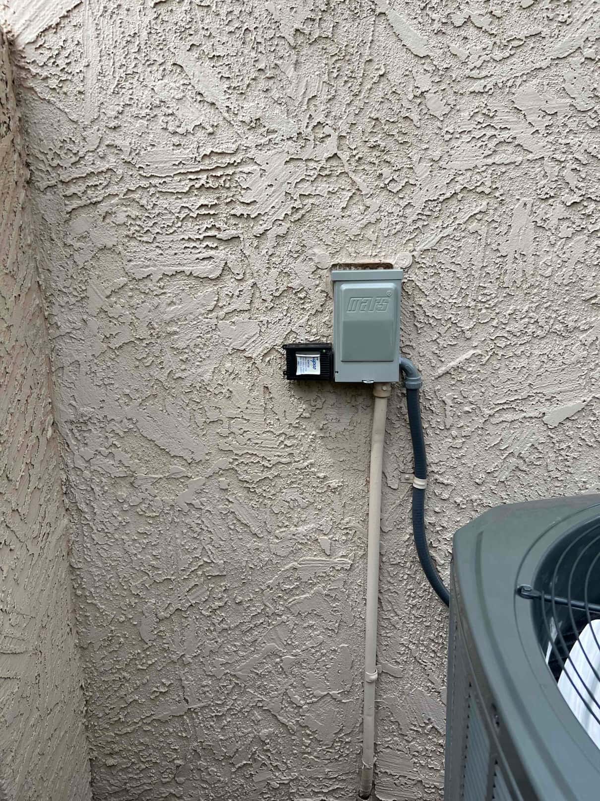 Outdoor service disconnect and electrical conduit installed beside the new condenser on a textured exterior wall.