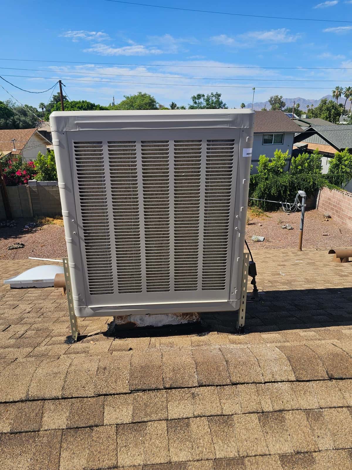 Rooftop evaporative cooler newly installed on support brackets, secured to shingles with sealed curb and visible electrical whip.