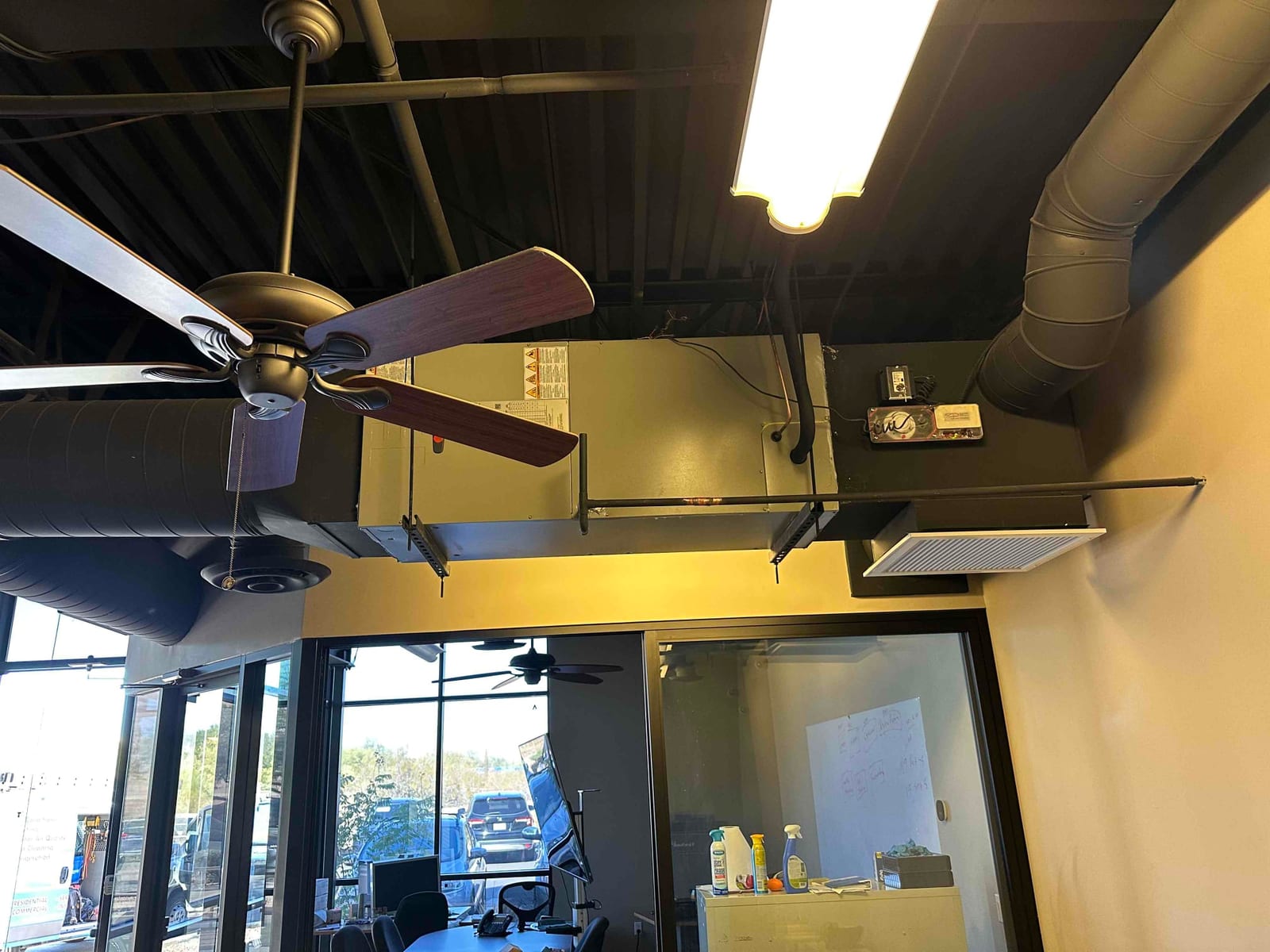 Suspended air handler with connected round ductwork and return grille installed over an office area with ceiling fans.