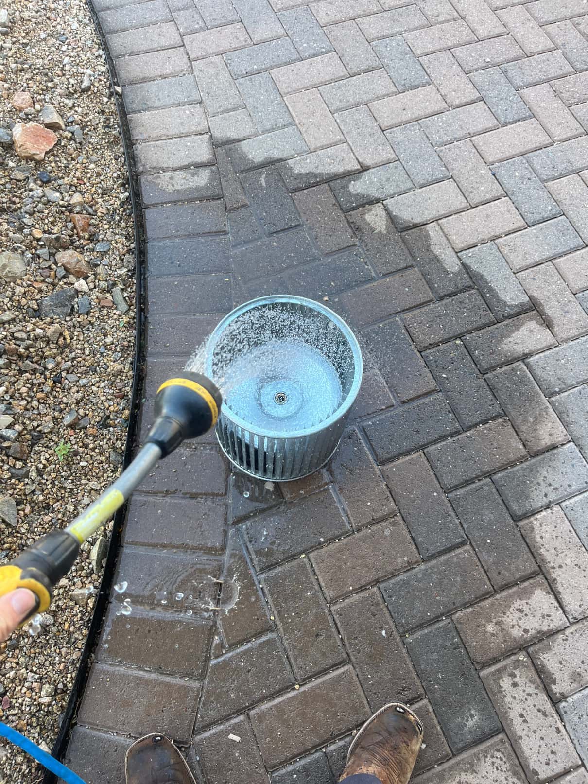 Technician rinses debris from a removed HVAC blower wheel with a hose on a paved surface.