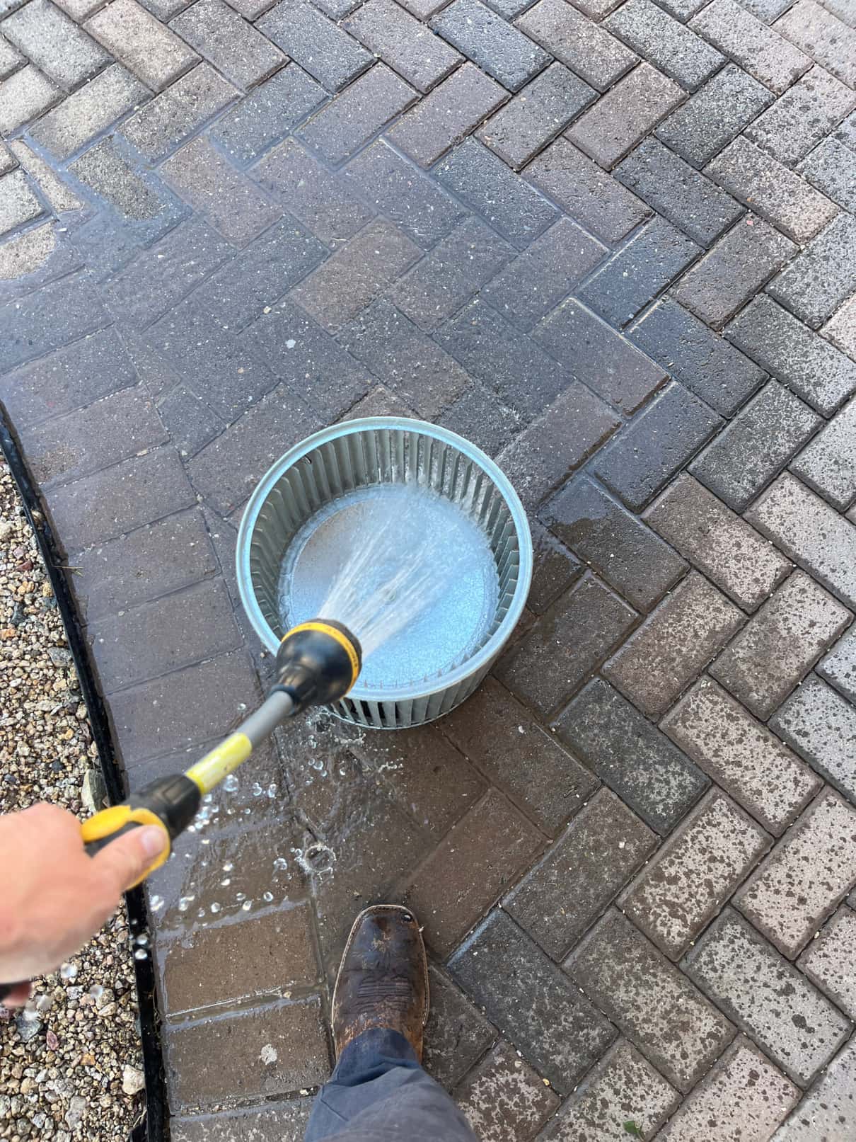 Technician rinses a metal blower wheel with a hose nozzle on a paved surface, removing debris from the fins.
