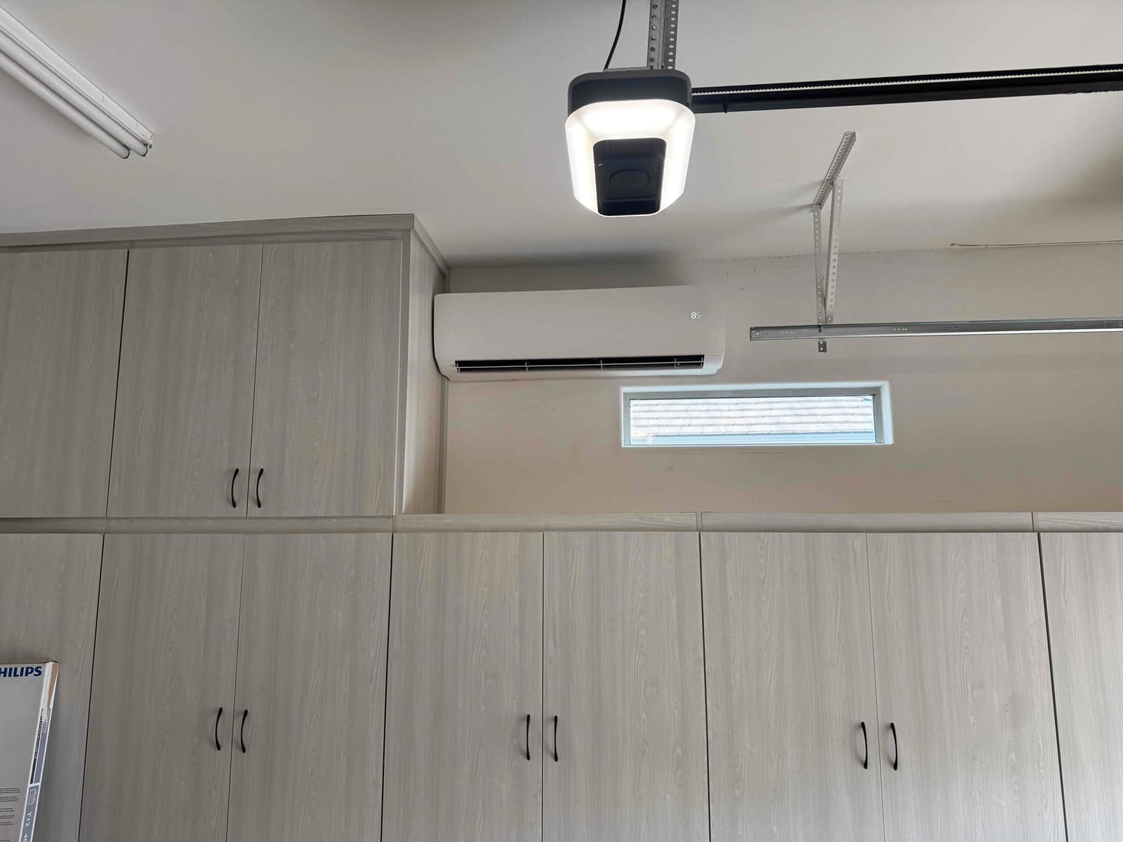 Wall-mounted mini-split indoor unit installed above built-in cabinets in a finished garage, positioned near a high window and ceiling tracks.