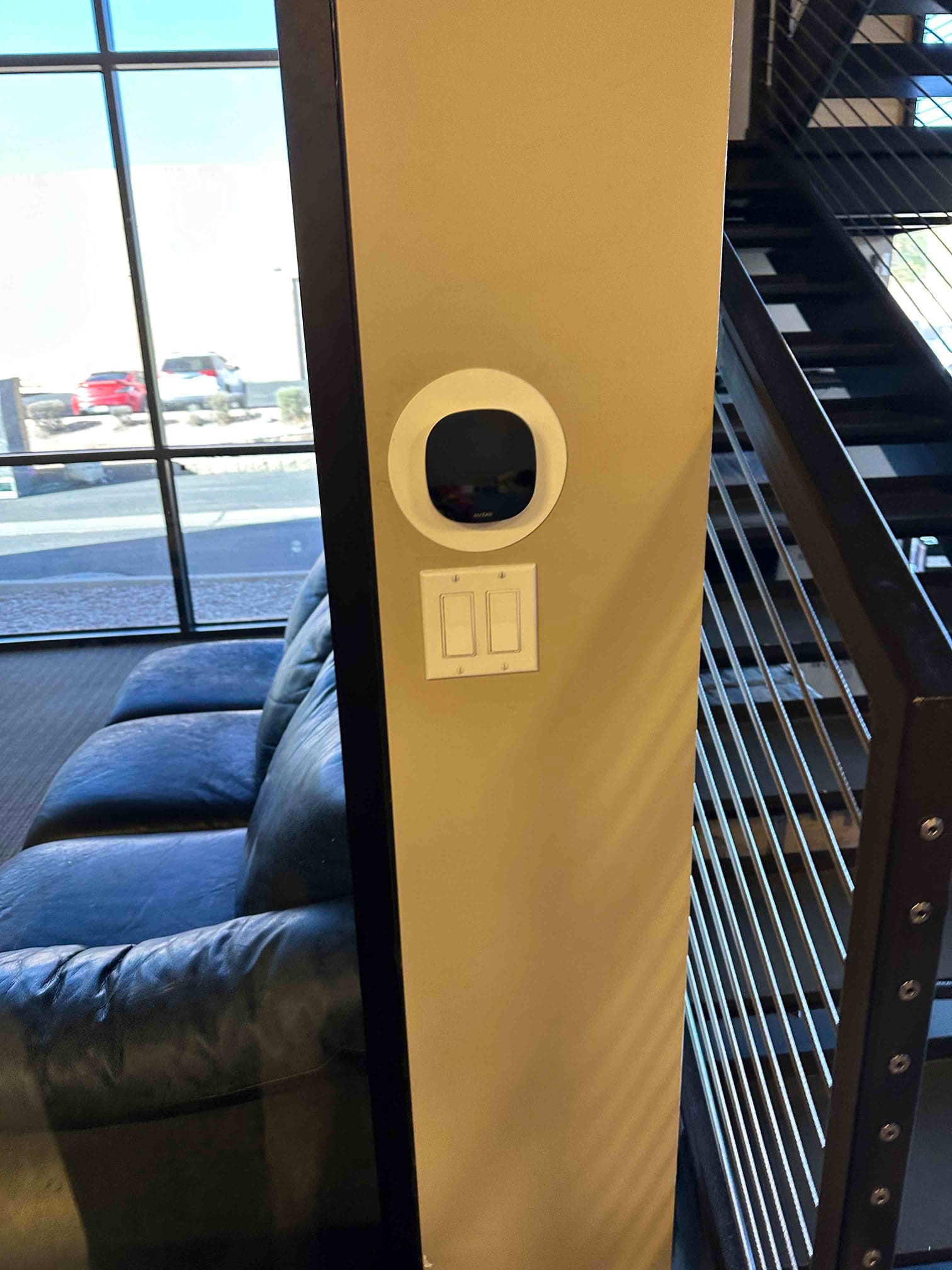 Wall-mounted smart thermostat backplate installed near a stair railing, with adjacent dual light switches on a finished interior wall.