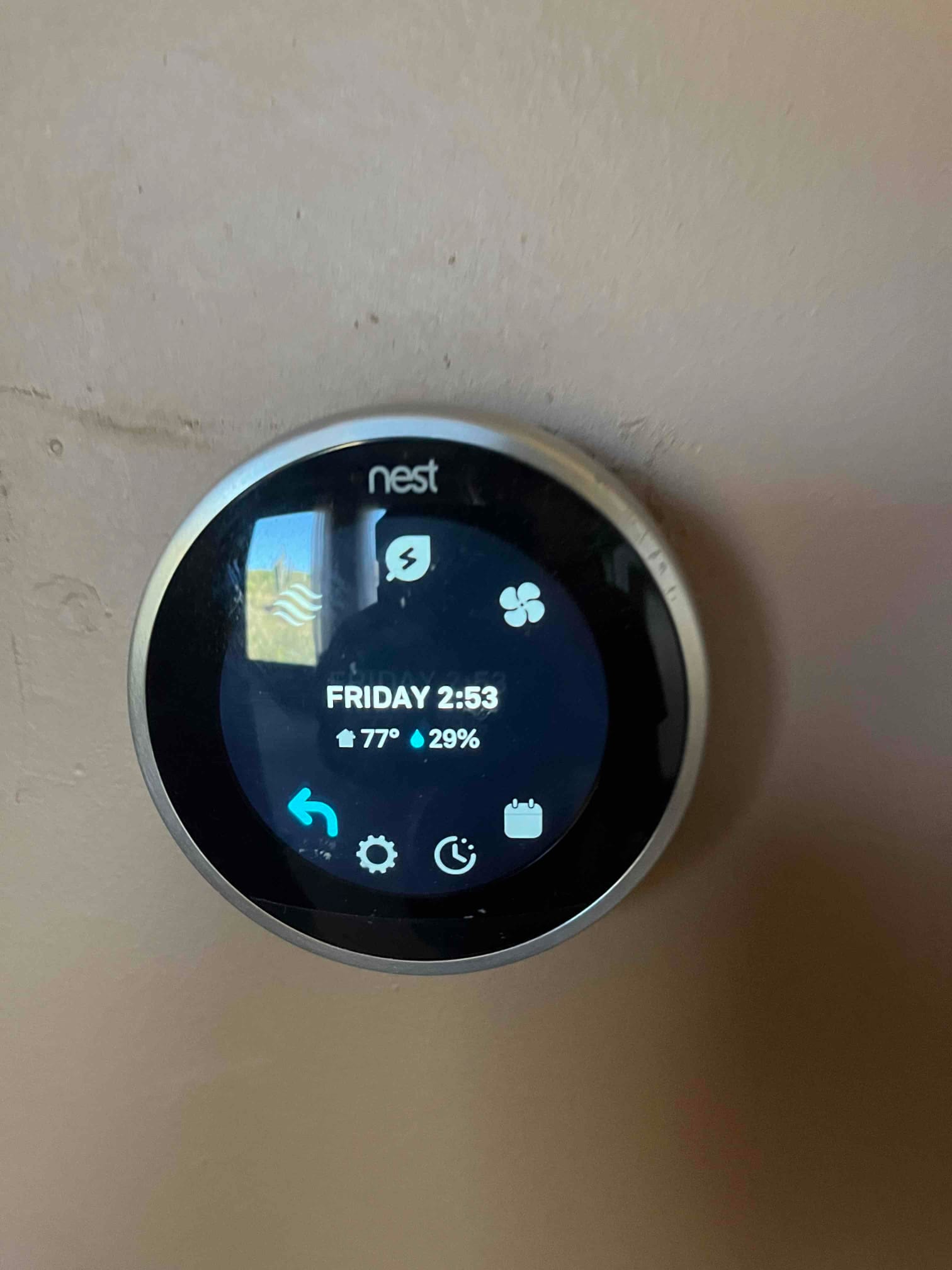 Wall-mounted smart thermostat displays time and indoor conditions, showing 77 degrees and 29 percent relative humidity.