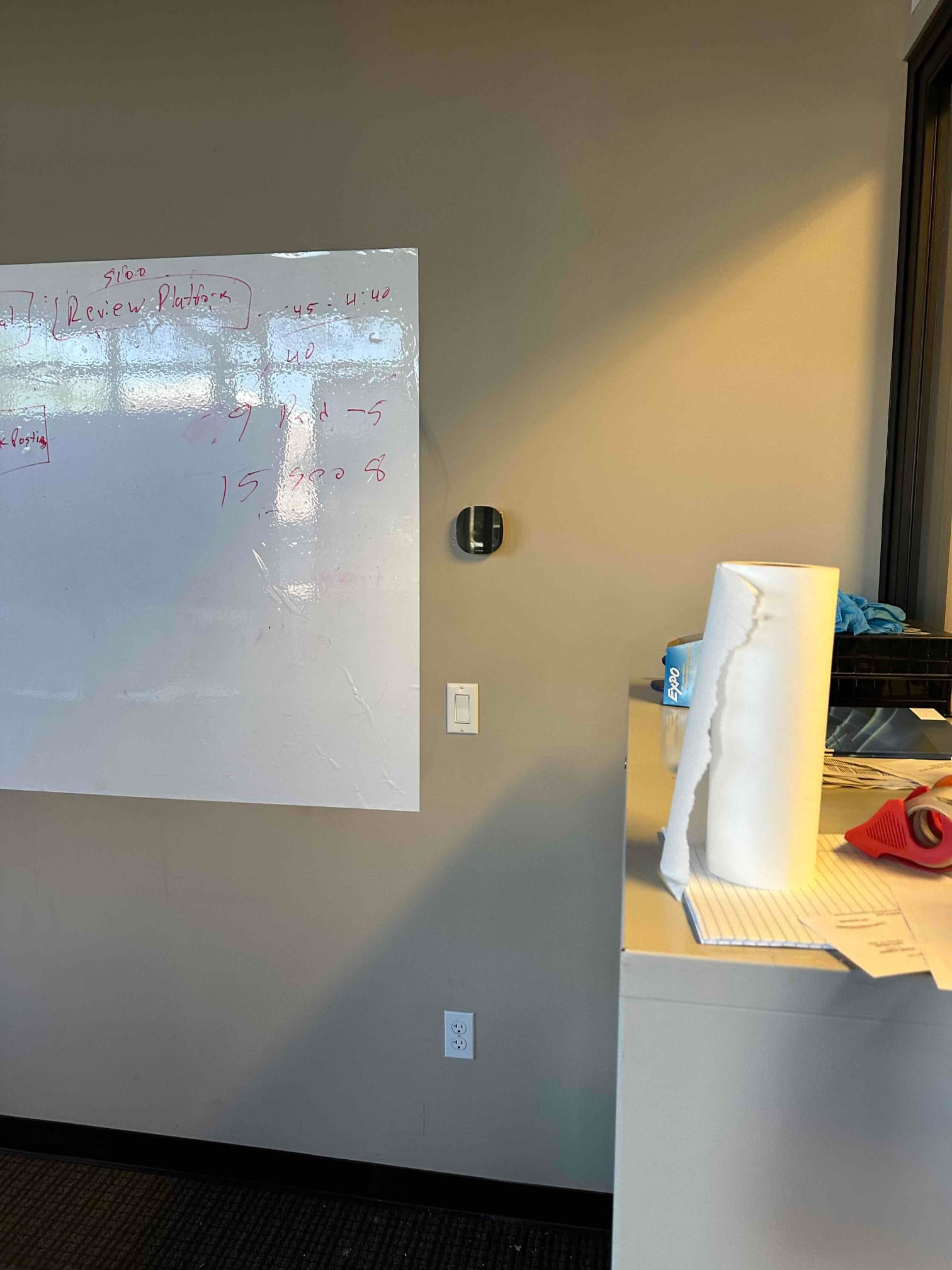 Wall-mounted smart thermostat installed in an office space near a whiteboard, with nearby light switch and outlet visible.