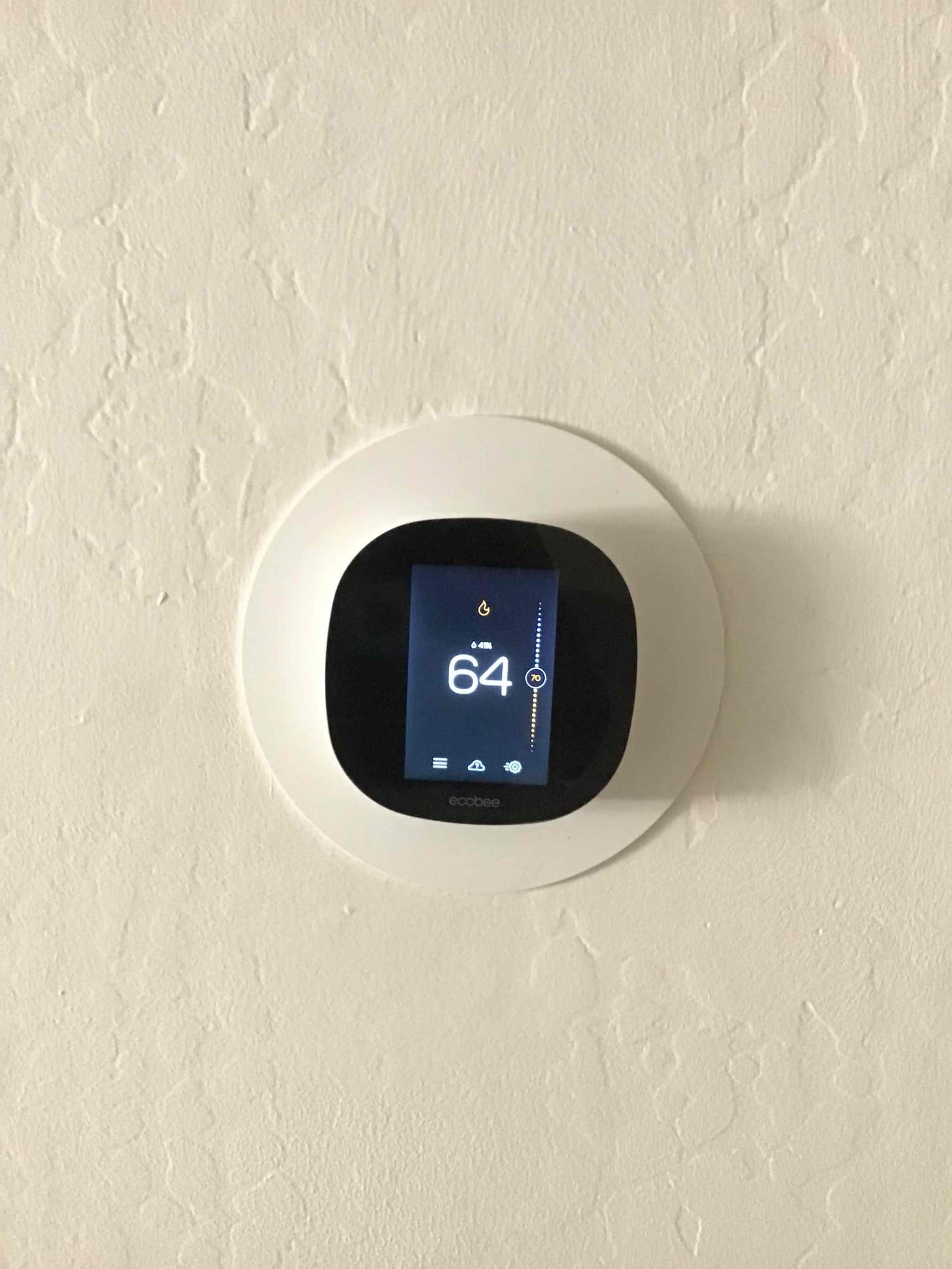 Wall-mounted smart thermostat installed with trim plate, screen displaying room temperature of 64 degrees and setpoint indicator.