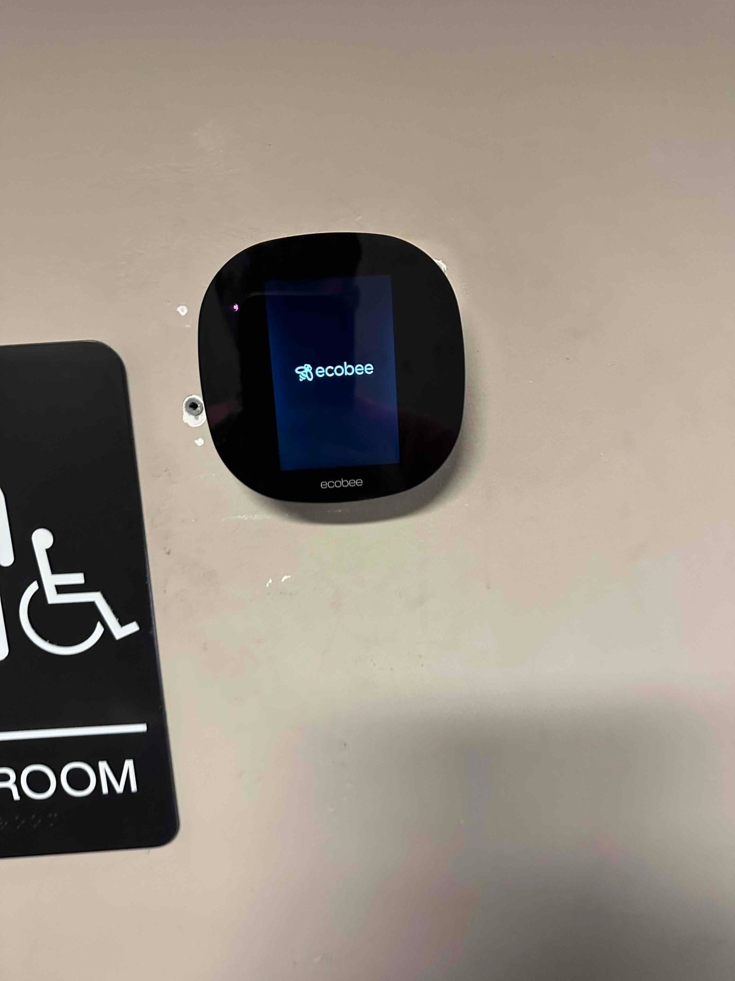 Wall-mounted smart thermostat powers on after installation, displaying its startup screen adjacent to a room sign.