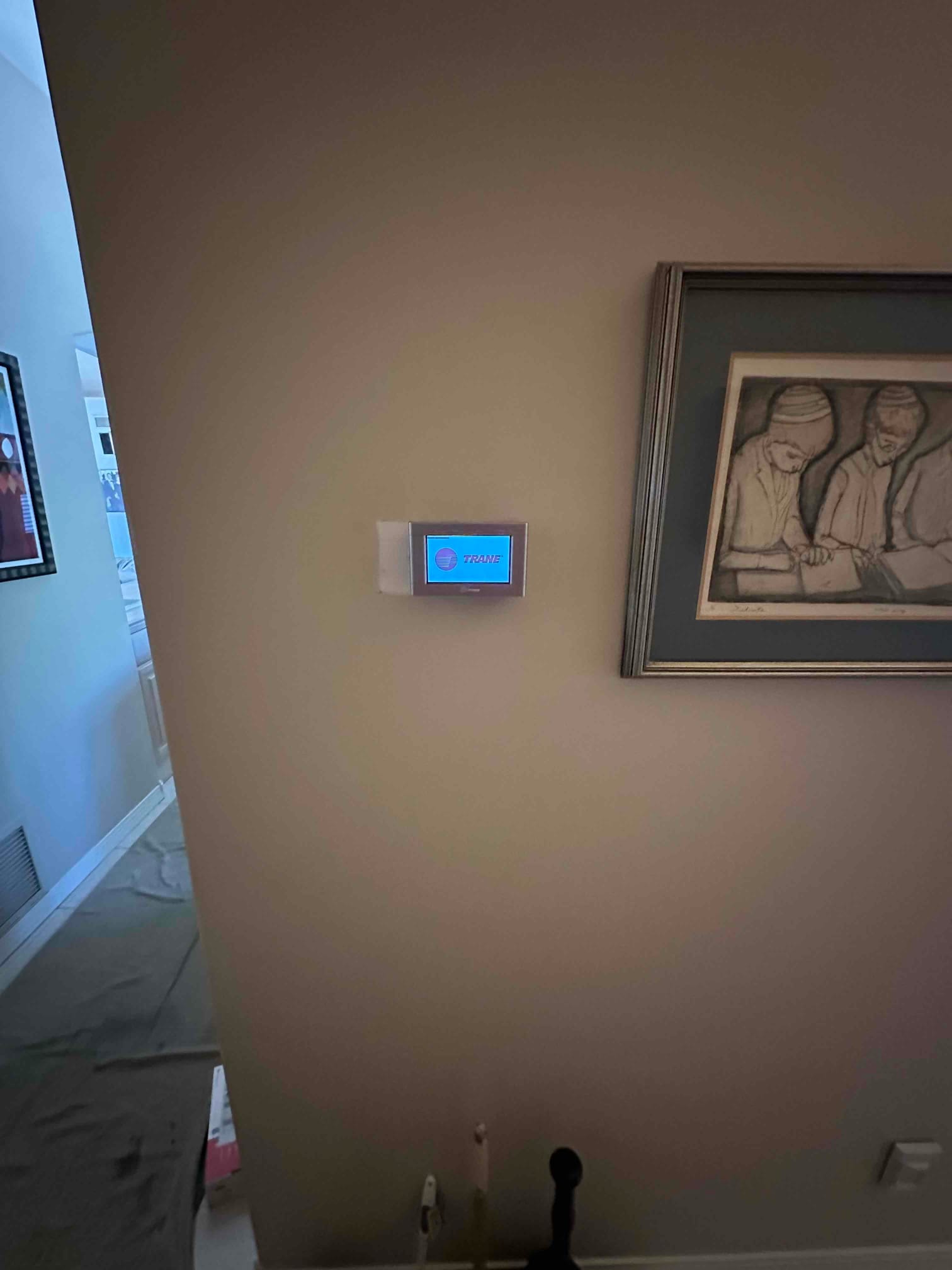 Wall-mounted thermostat installed and powered on in hallway adjacent to return grille and protective floor covering.
