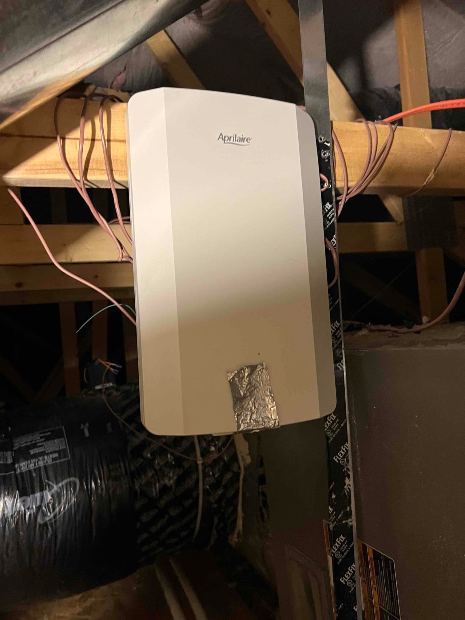 White equipment housing mounted on the duct plenum in the attic, with multiple low-voltage wires and foil tape.