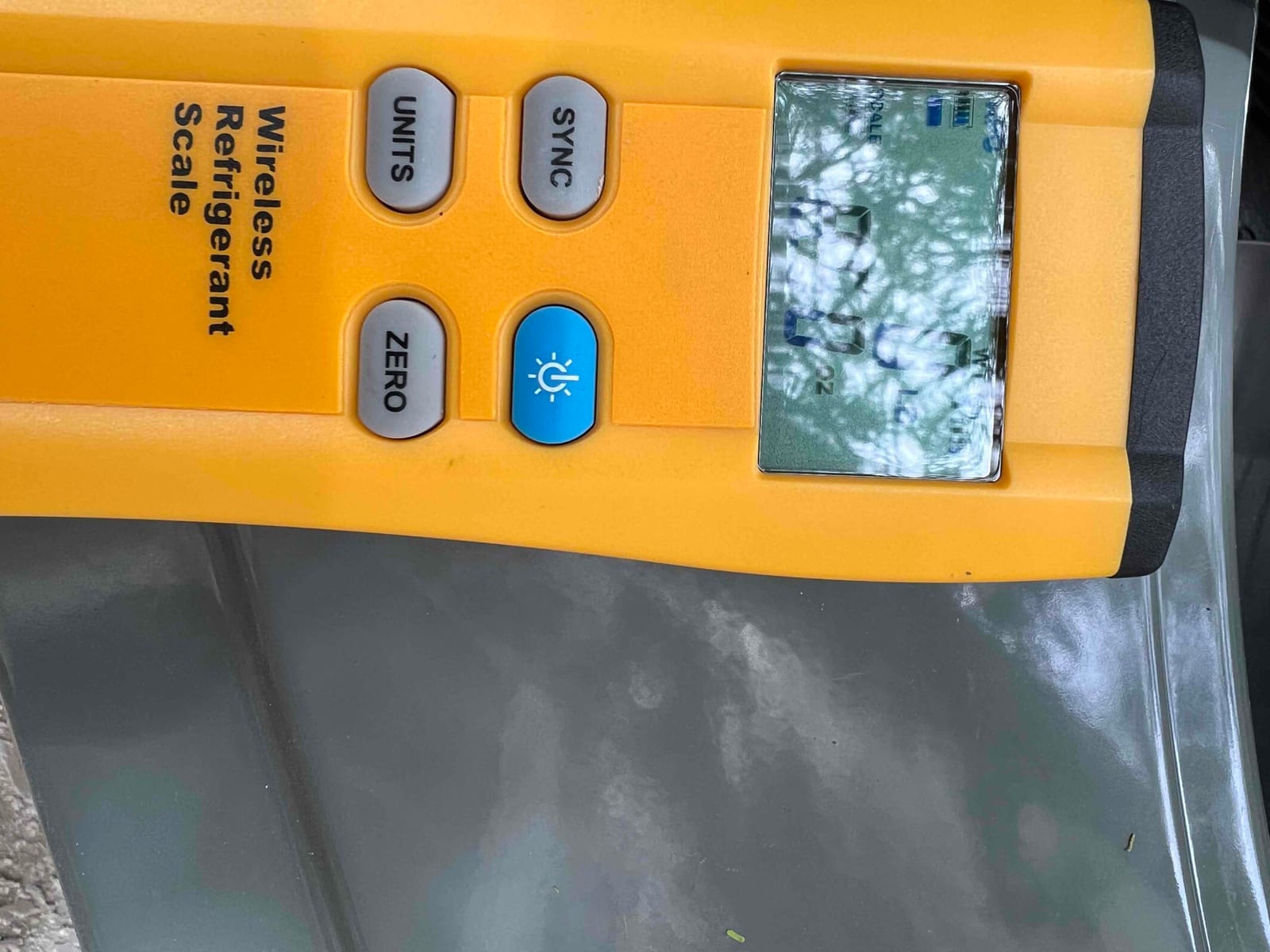 Wireless refrigerant scale sits on the outdoor unit during charging, with screen obscured by glare and unreadable.
