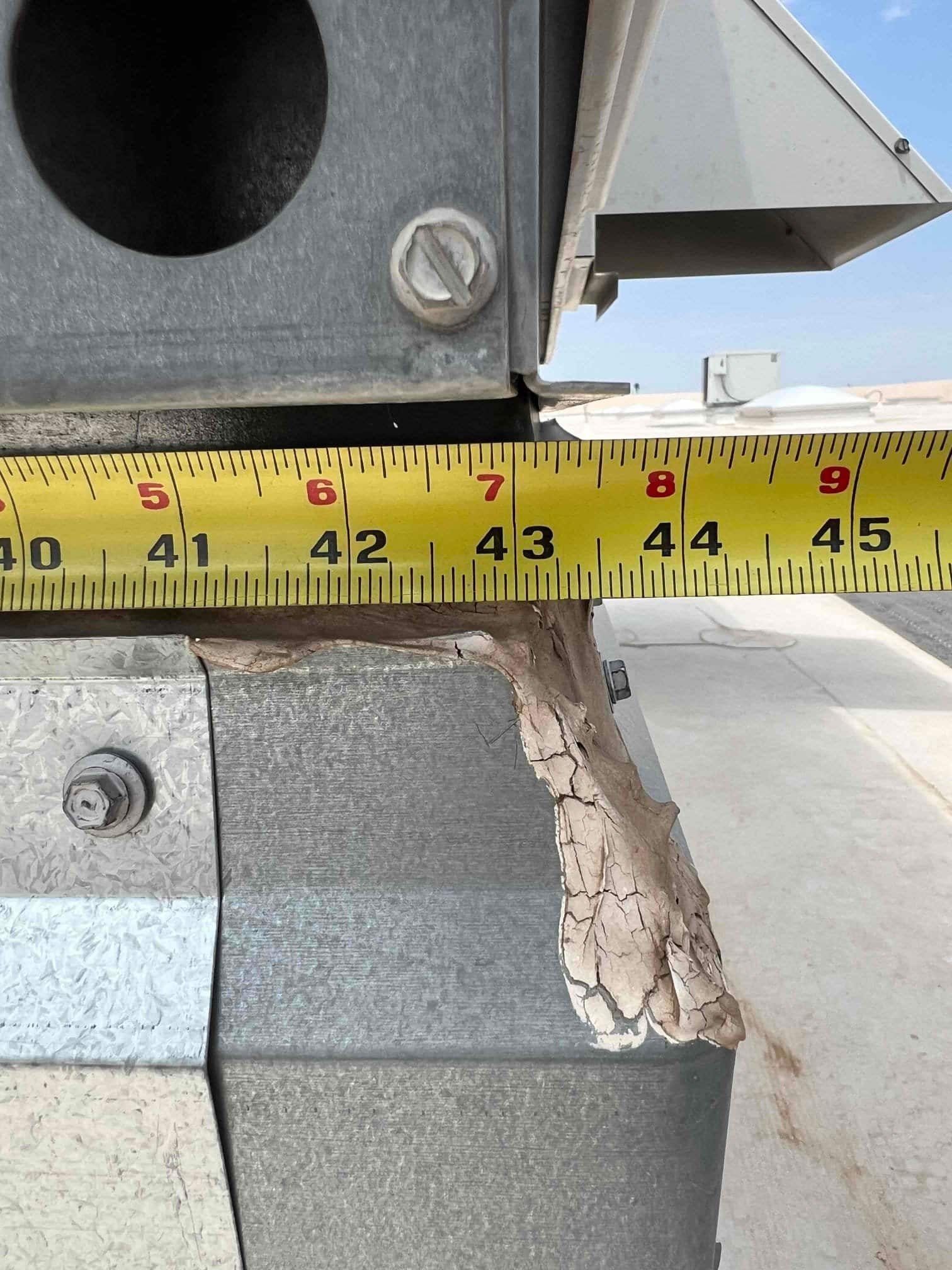 Close-up of rooftop HVAC duct curb with cracked and peeling sealant, measured across with a tape showing approximately 6 to 9 inches.
