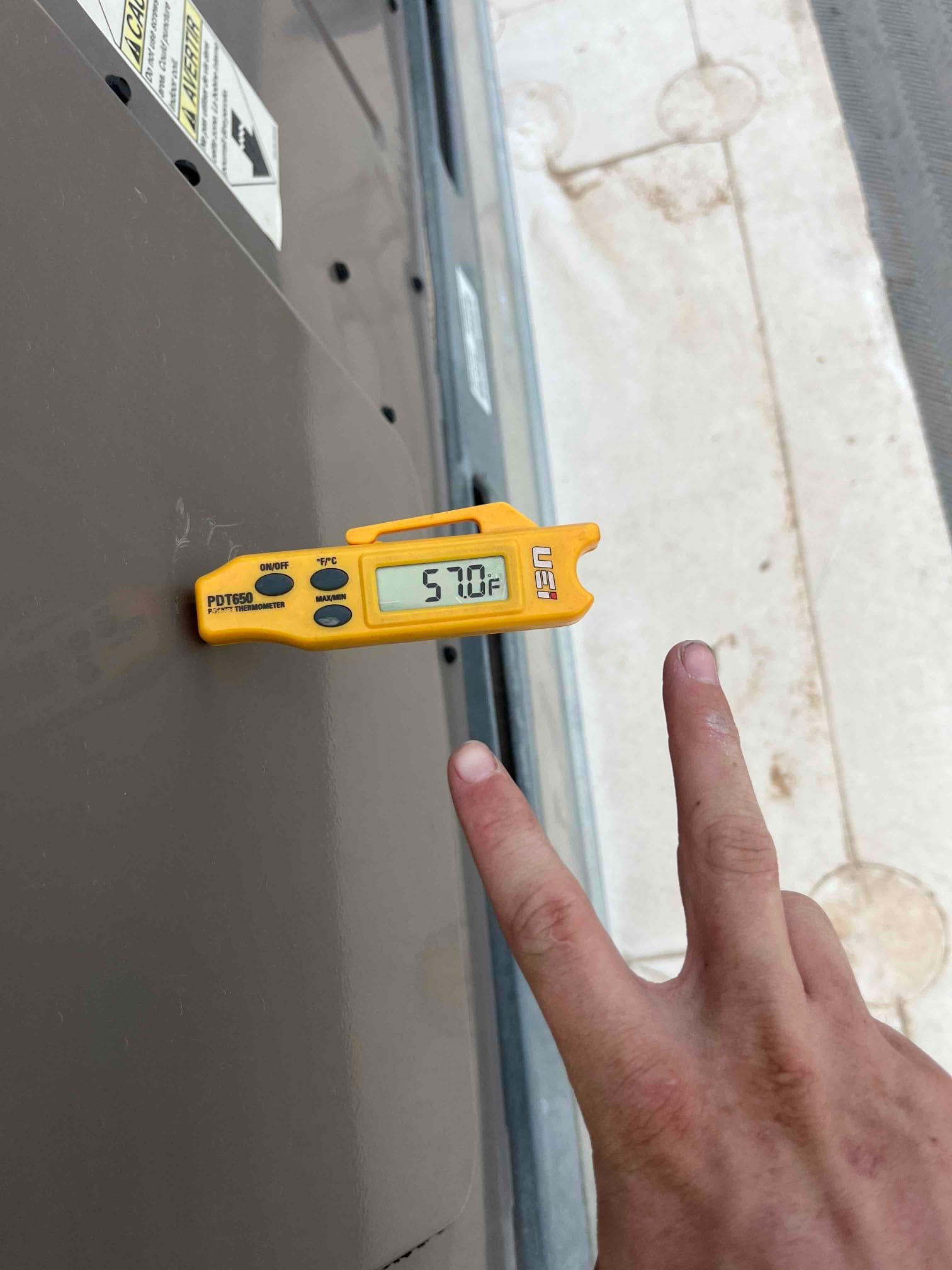 Digital thermometer shows 57.0 °F surface temperature on the HVAC cabinet during air conditioning repair.