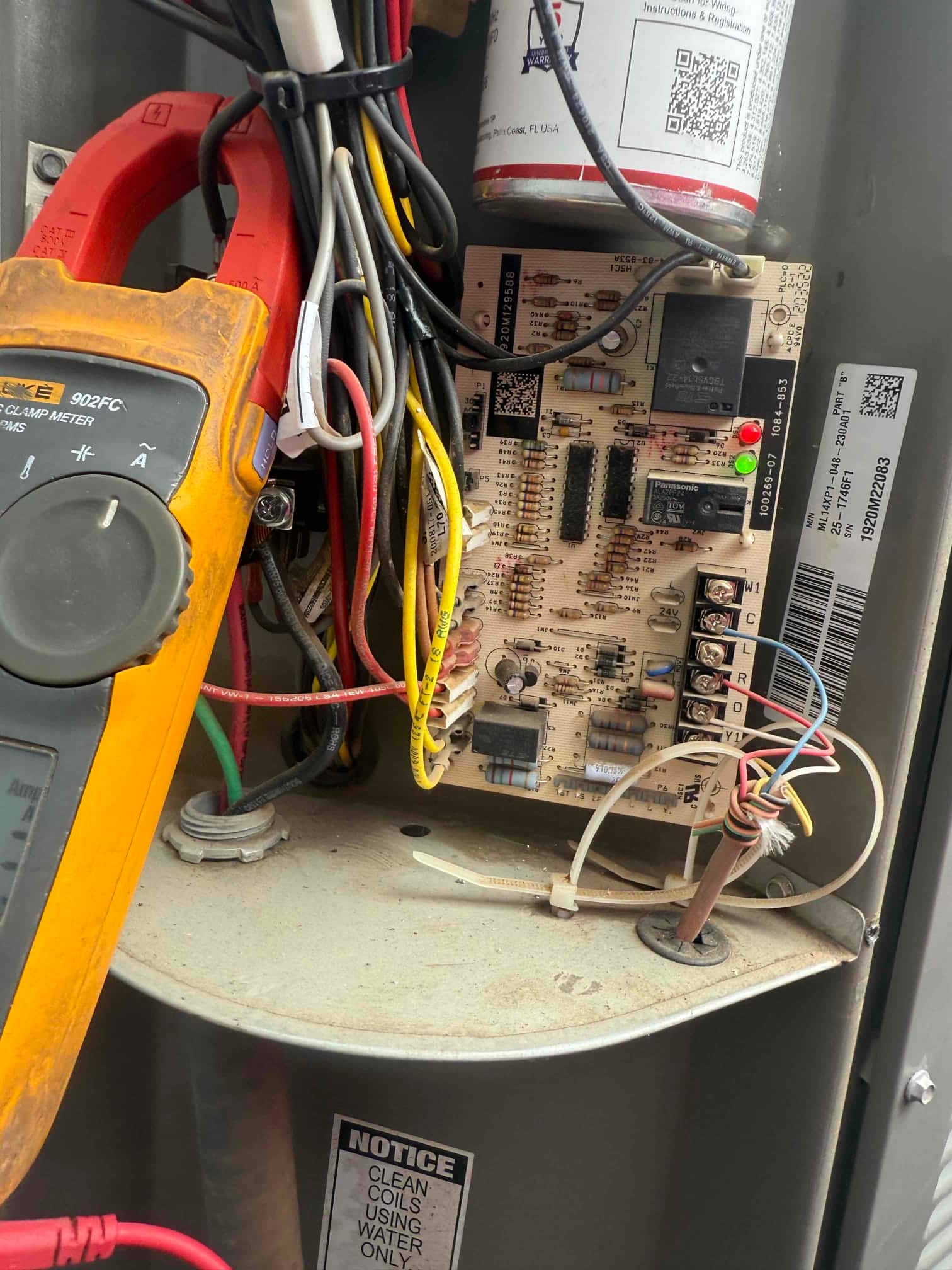 Furnace control board exposed with wiring, installed dual-run capacitor above, and clamp meter positioned nearby for diagnostics.