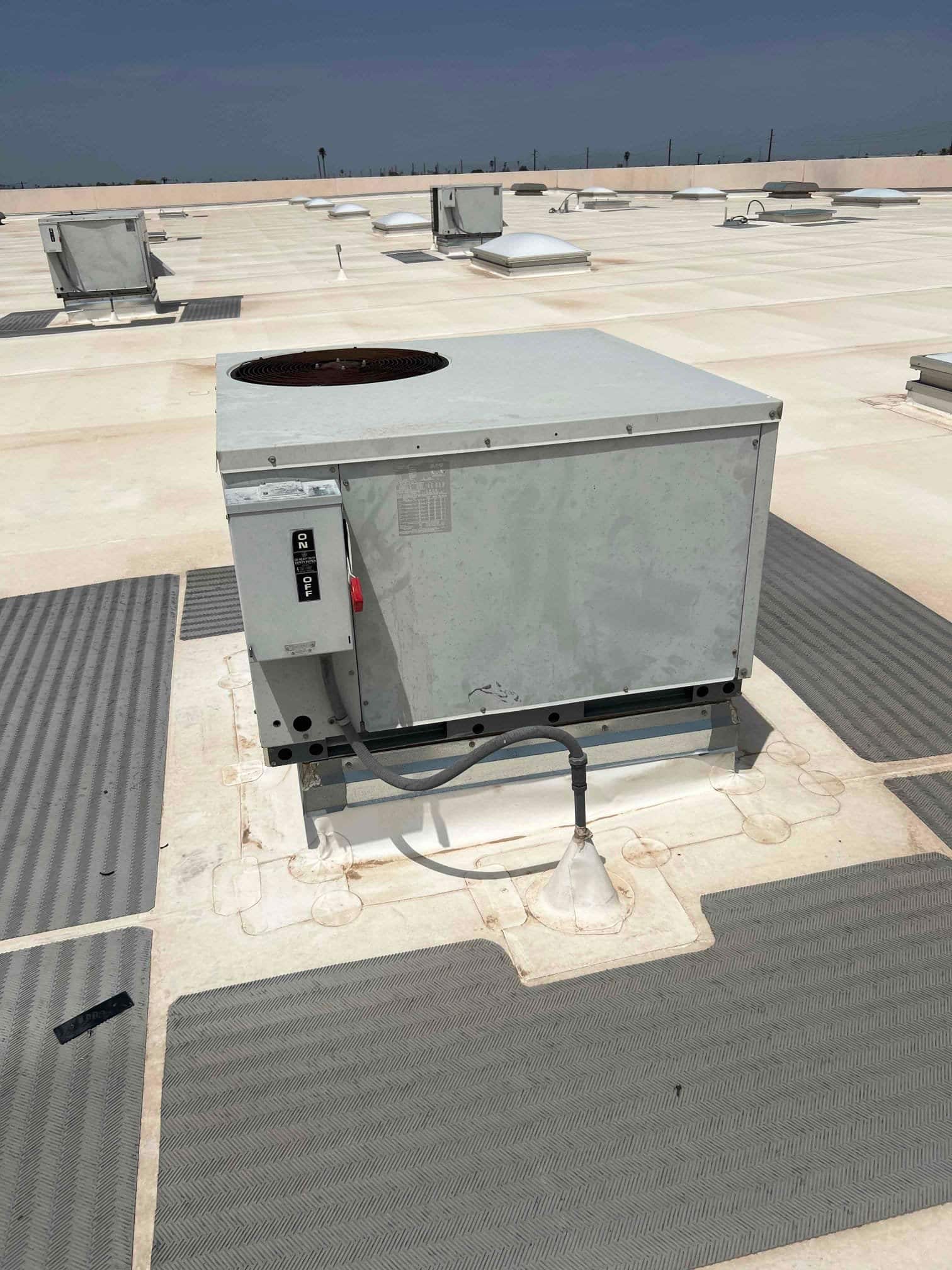 Rooftop air conditioning condenser unit with external disconnect and flexible conduit, showing weathered housing on a flat commercial roof.